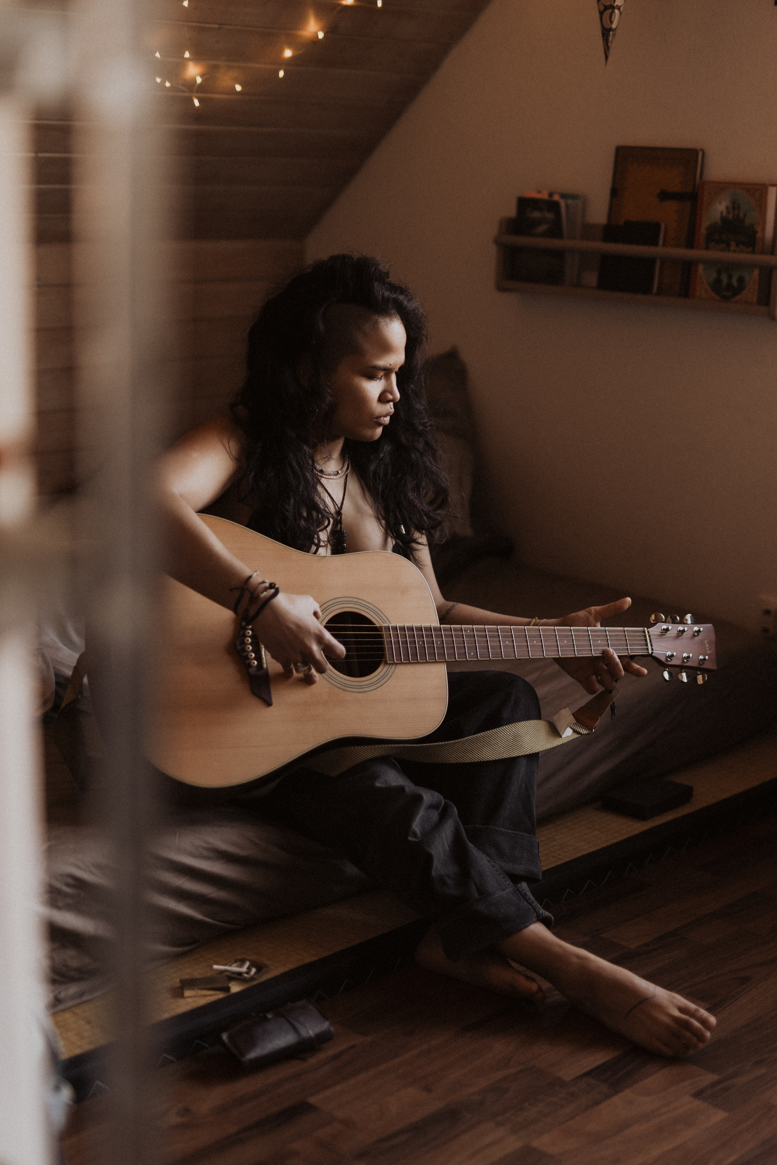 Eine Person mit langen Haaren sitzt auf einem Bett in einem gemütlichen, schwach beleuchteten Raum und spielt Akustikgitarre. Von der Decke hängen Lichterketten, im Hintergrund stehen Bücher und Dekoartikel auf einem Regal. Die Atmosphäre ist warm und entspannt und schafft eine ideale Kulisse für ein intimes Boudoir-Fotoshooting zu Hause.