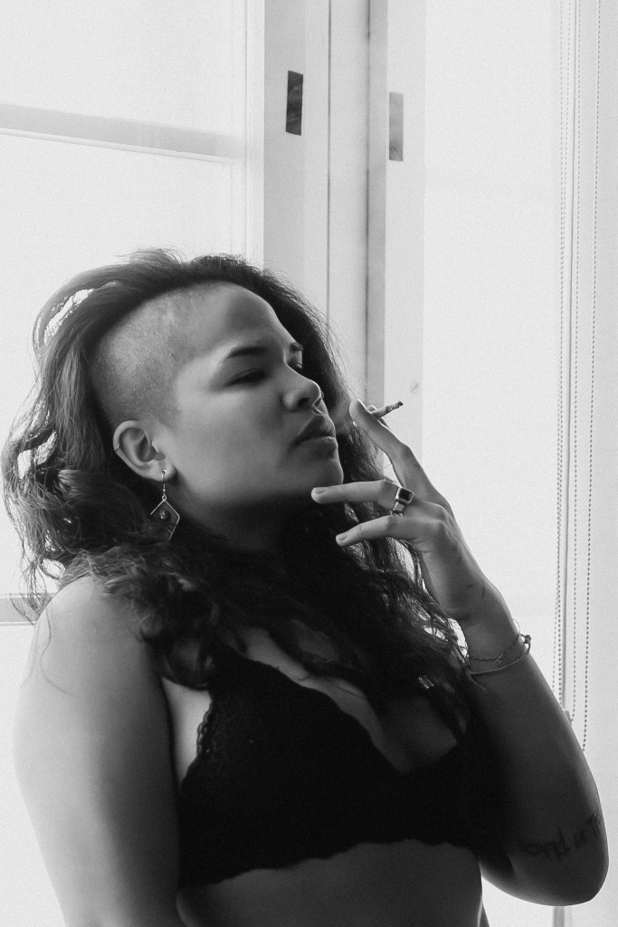 Eine Frau mit langen, lockigen Haaren und einer rasierten Seite trägt einen schwarzen Spitzen-BH und diversen Schmuck. Sie raucht eine Zigarette vor einem Fenster zu Hause, ihr Gesichtsausdruck ist ruhig und entspannt. Das von der Fotografin wunderschön aufgenommene monochrome Porträt verleiht der Szene einen Hauch zeitloser Eleganz.