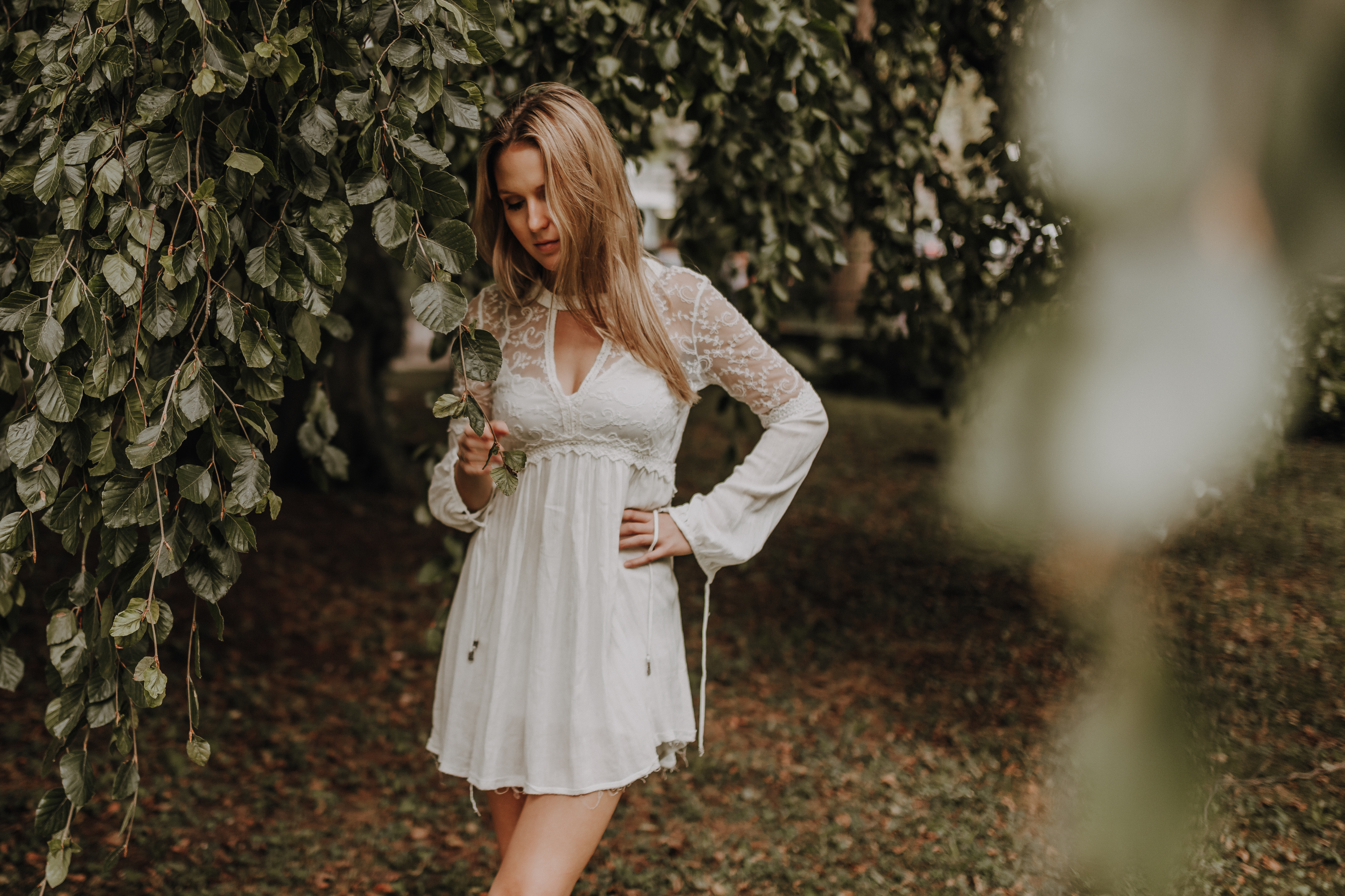 Eine blonde Frau steht unter einer Trauerbuche. Sie trägt ein weisses Spitzenkleid, welches einen tollen contrast zum dunklen Grün der Blätter bildet auf dieser magischen Portraitaufnahme von Tasha Ender Photography.