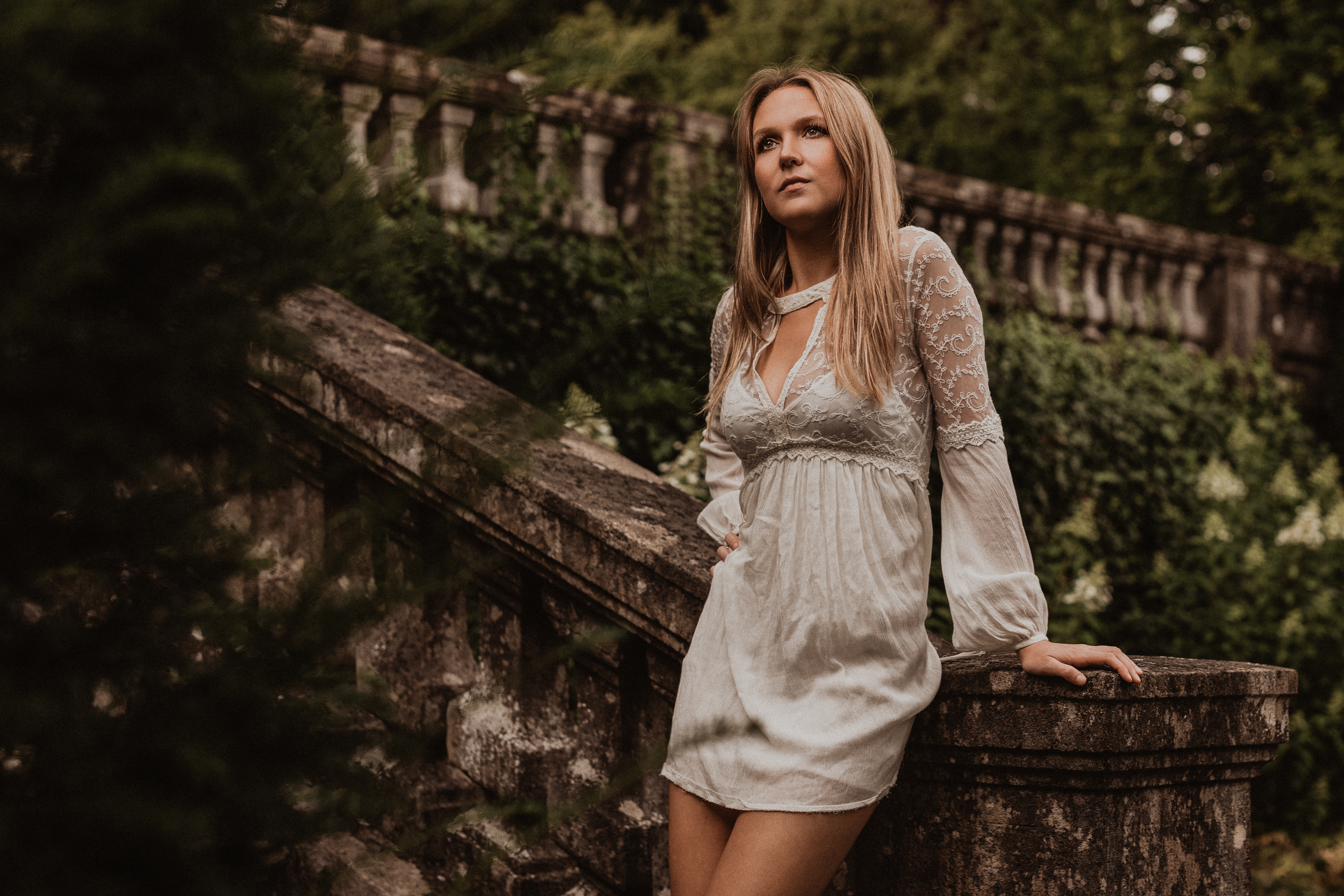 Eine blonde Frau in einem weissen Spitzenkleid lehnt sich entspannt gegen eine steinerne Treppe. Der verschwommene Hintergrund lässt erahnen, dass das Bild im Villa Boveri Park in Baden aufgenommen wurde.