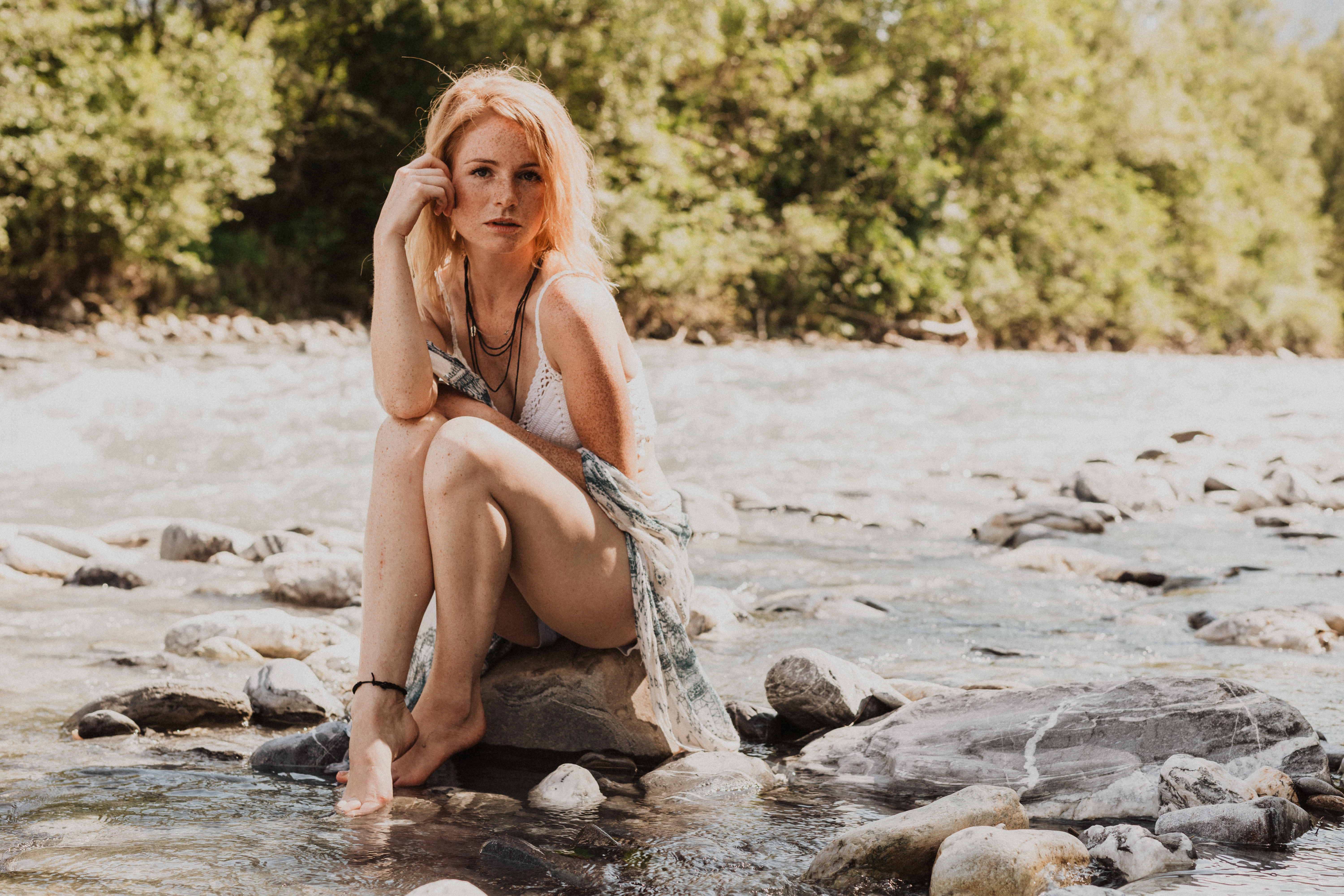 Eine Frau mit langen roten Haaren posiert sinnlich für ein Portrait während eines Fotoshootings. Sie sitzt auf einem Stein im Wasser und schaut in die Kamera der Fotografin für Frauen.