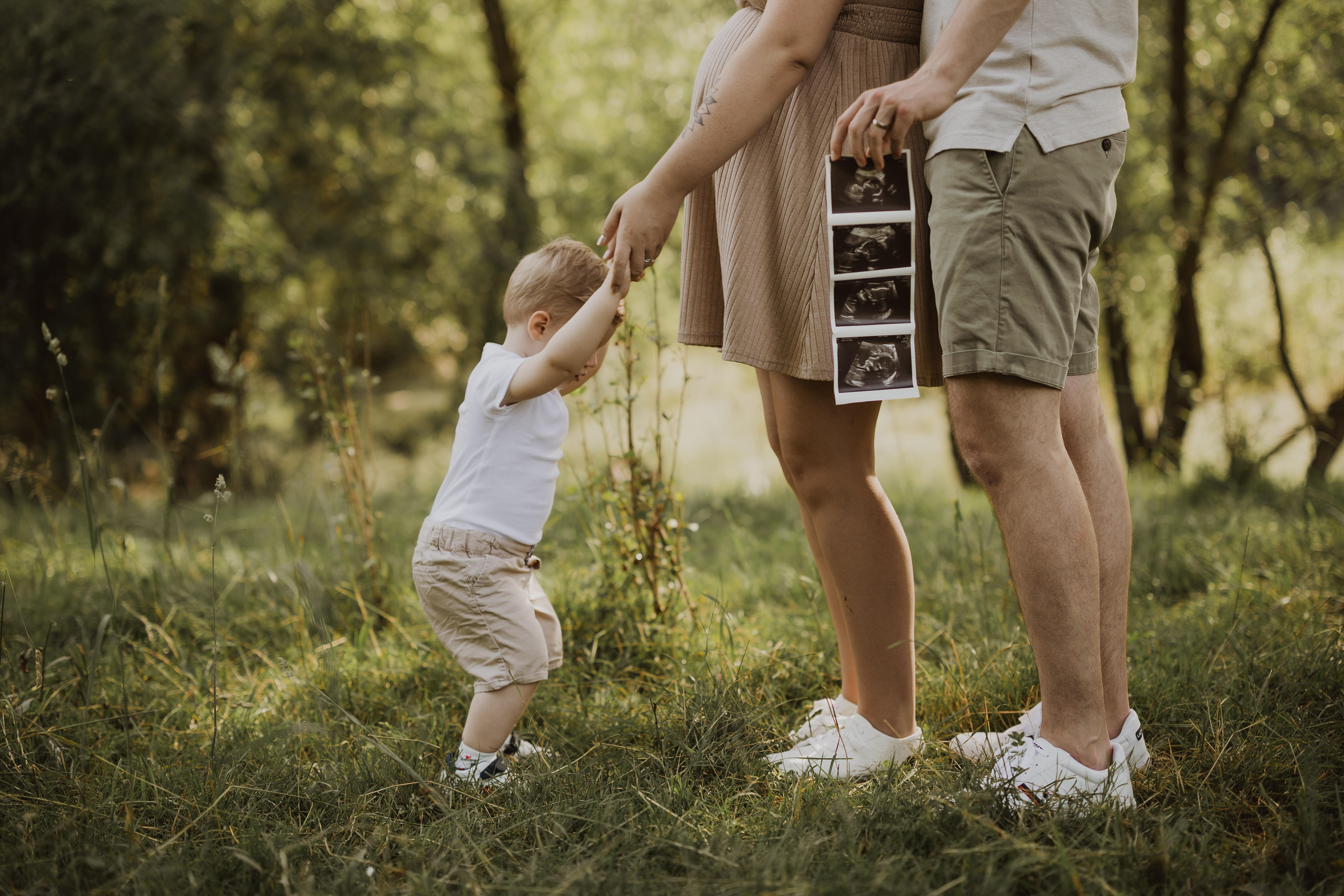Ein junges Paar steht draußen auf einer Wiese und hält sein Kleinkind an der Hand. Die Frau hält Ultraschallbilder in einer Hand, während der Mann neben ihr steht. Beide Erwachsenen tragen lässige Sommerkleidung und das Kleinkind streckt sich nach oben, um die Hand der Frau während ihres Babybauch-Fotoshootings zu berühren.