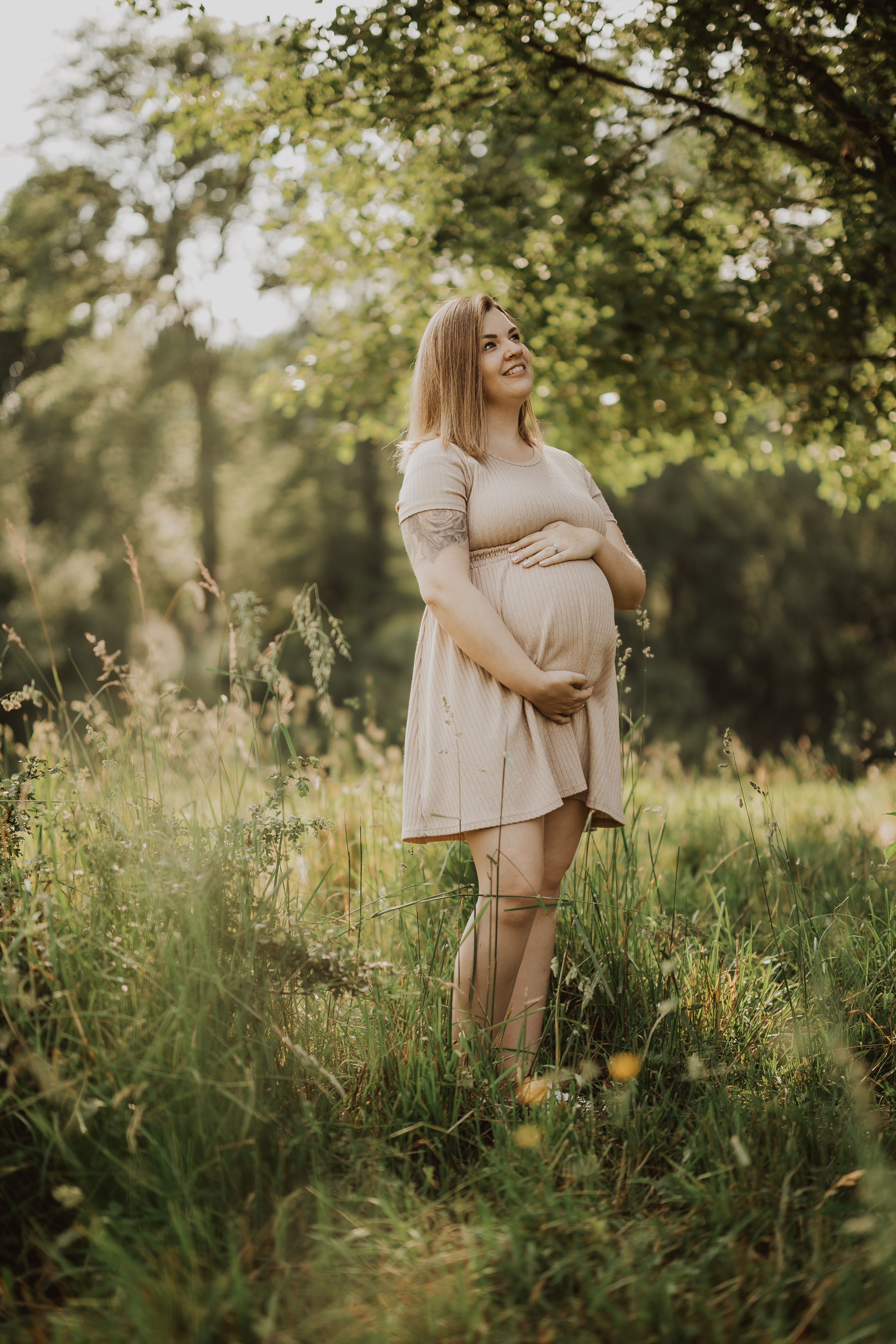 Eine schwangere Frau in einem hellen Kleid steht auf einer sonnenbeschienenen Wiese, umgeben von hohem Gras und Bäumen. Sie hält ihren Bauch mit beiden Händen und blickt mit heiterem Gesichtsausdruck nach oben. Der Hintergrund ist üppig grün und schafft eine friedliche Atmosphäre, perfekt für ein Babybauch-Fotoshooting.