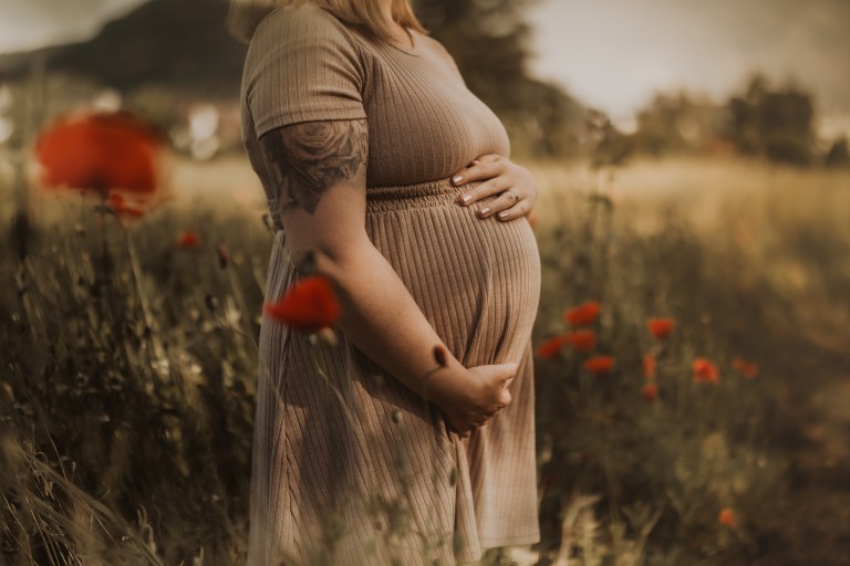 Eine schwangere Frau in einem braunen Rippenkleid steht in einem üppigen Feld aus Wildblumen und hält ihren Bauch mit beiden Händen. Sie hat blonde Haare und ein Tattoo auf dem linken Oberarm. Der Hintergrund ist sanft verschwommen mit roten Mohnblumen und Grün. Es ist eine perfekte Kulisse für ein Schwangerschaftsshooting in der Natur.