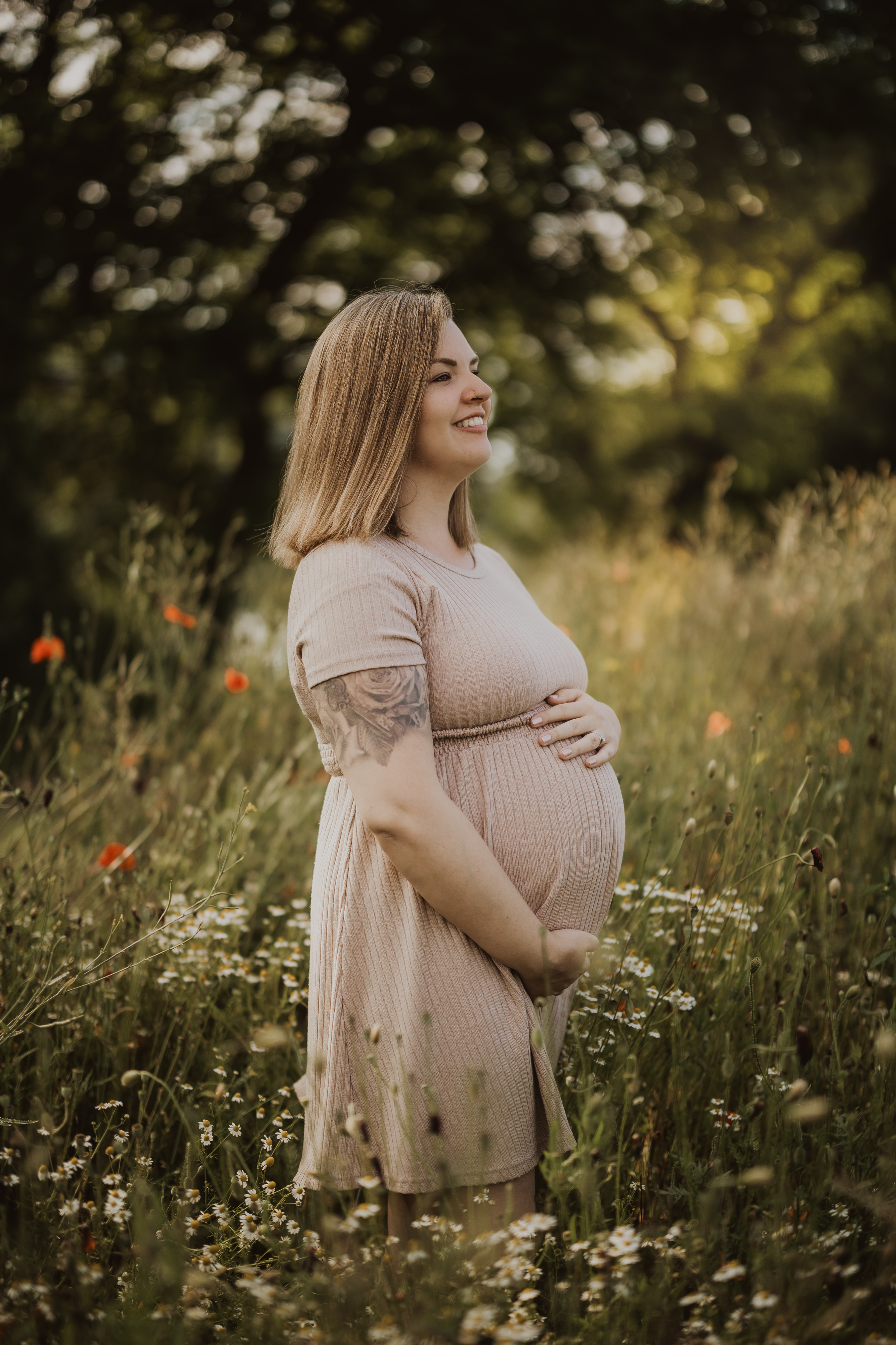 Eine schwangere Frau steht in einem Feld voller Wildblumen und hält ihren Bauch in den Händen. Sie trägt ein hellrosa Kleid und hat schulterlanges Haar. Mit einem natürlichen Lächeln blickt sie in die Ferne, umgeben von Grün und Blumen – eine idyllische Szene, perfekt für ein Babybauch-Fotoshooting.