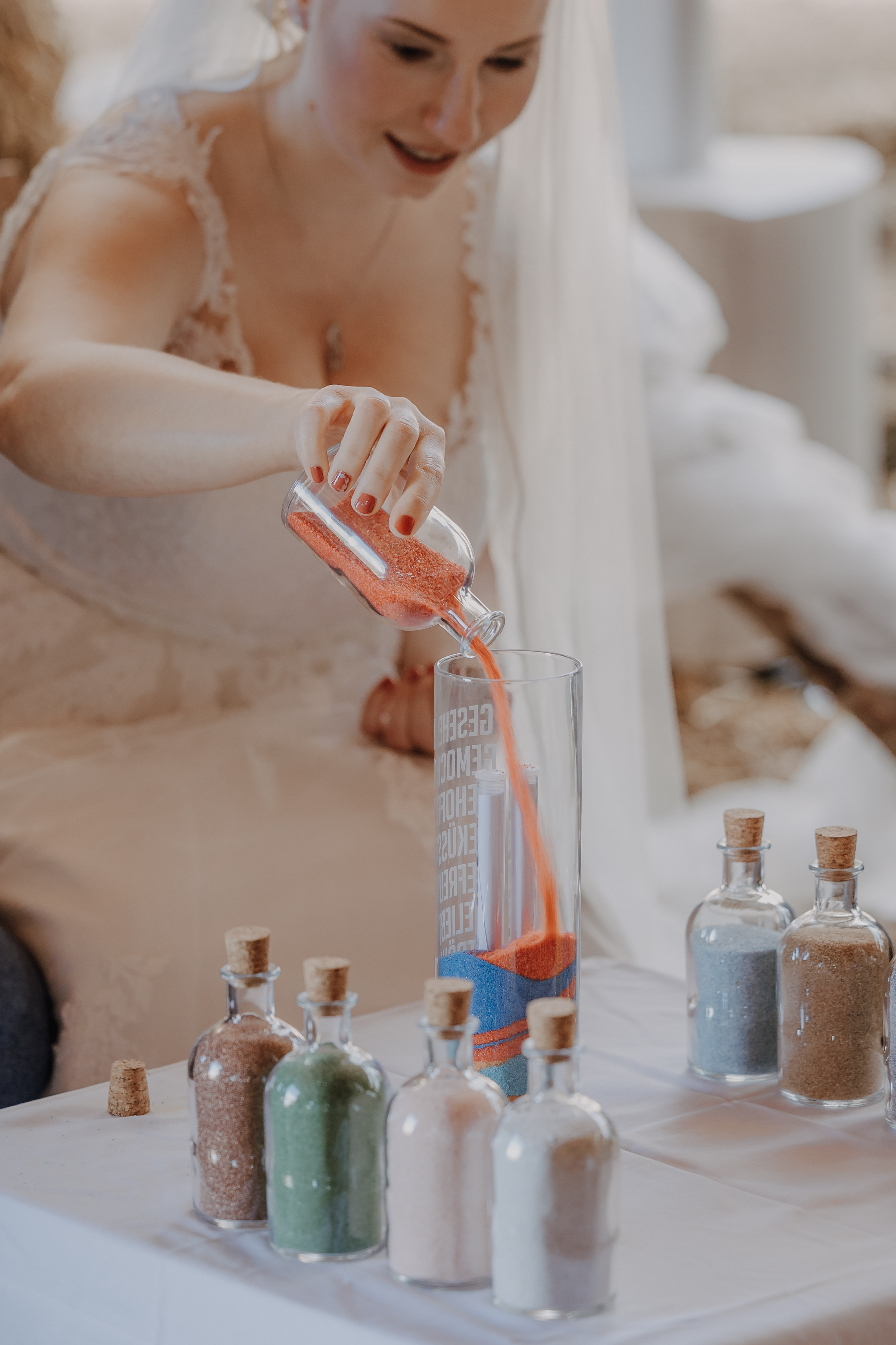 Eine Braut im Hochzeitskleid, aufgenommen von Tasha Ender Photographys Hochzeitsfotografie-Service, führt eine umweltbewusste Sandzeremonie durch. Sie schüttet roten Sand in eine hohe Glasvase, in der sich bereits blauer Sand befindet. Auf dem Tisch vor ihr stehen mehrere kleine Flaschen mit umweltfreundlichem farbigem Sand.