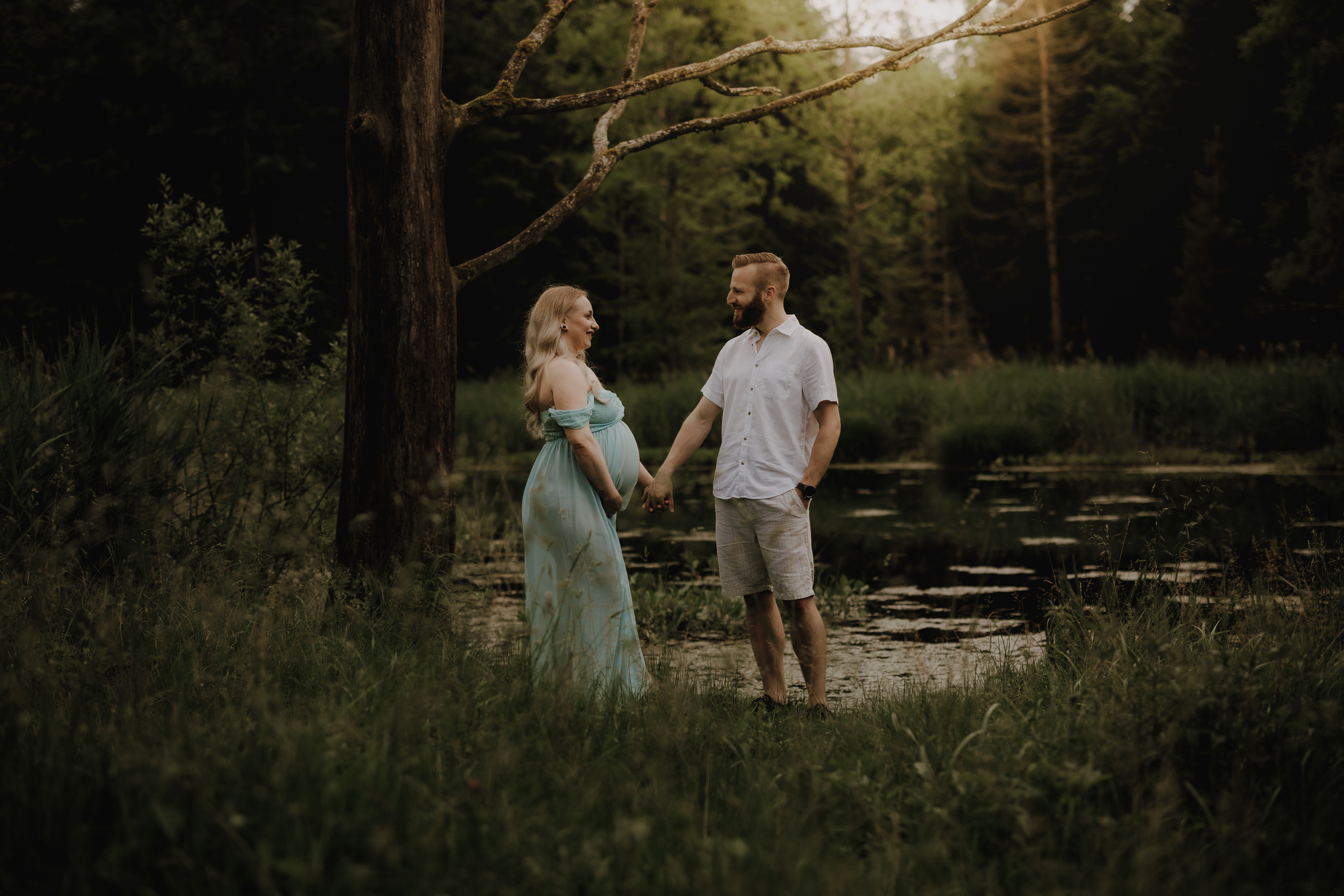 Eine schwangere Frau in einem fließenden blauen Kleid steht Händchen haltend mit einem Mann in weißem Hemd und Shorts an einem ruhigen Waldteich. Sie sind umgeben von üppigem Grün und hohen Bäumen, getaucht in weiches, warmes Licht – ein idyllischer Moment, aufgenommen während ihres Schwangerschafts-Fotoshootings.
