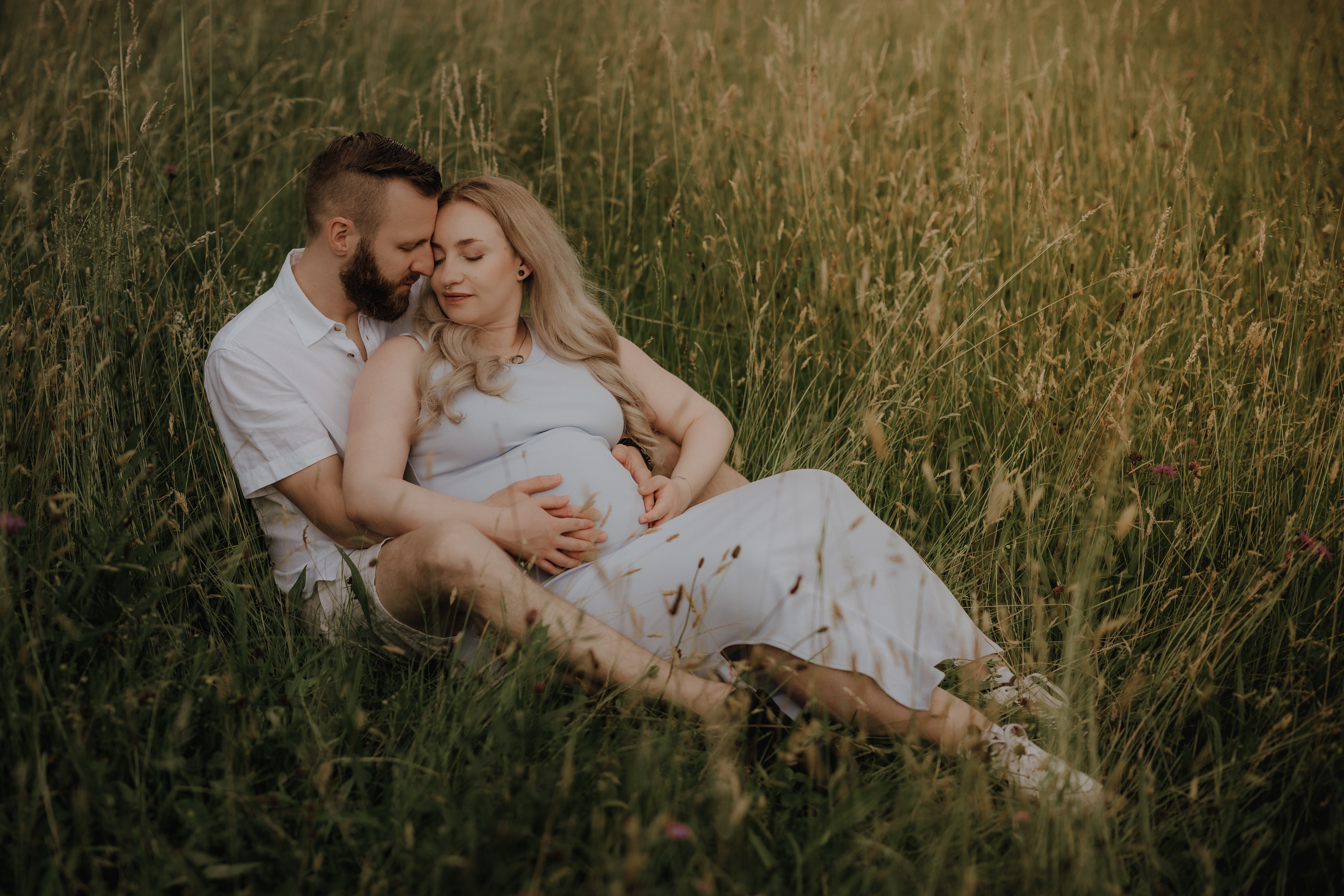 Ein Paar sitzt in einem Feld mit hohem Gras, der Mann sitzt hinter der Frau und umarmt sie. Beide tragen helle Kleidung. Die Frau ist schwanger und hat ihre Hände auf dem Bauch ihres Babys abgelegt. Beide wirken zufrieden und gelassen, perfekt eingefangen in diesem Schwangerschaftsshooting.