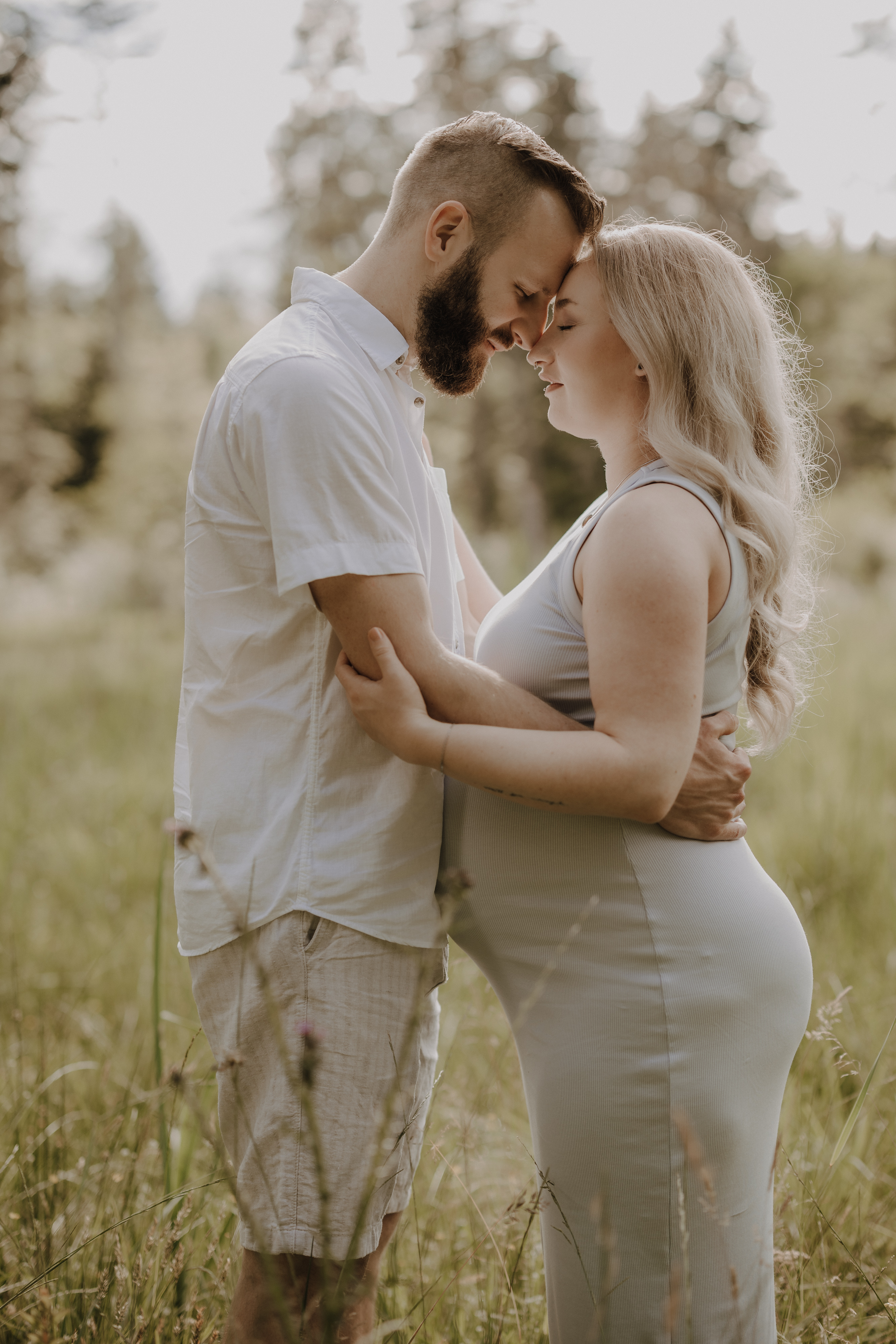 Ein Paar umarmt sich zärtlich auf einer sonnenbeschienenen Wiese während seines Schwangerschaftsshootings. Der Mann, bärtig und mit weißem Hemd und Shorts bekleidet, steht Stirn an Stirn mit der schwangeren Frau in ihrem hellen Kleid. Mit geschlossenen Augen sind sie von hohem Gras und Bäumen umgeben und fangen ihren freudigen Babybauch-Moment ein.