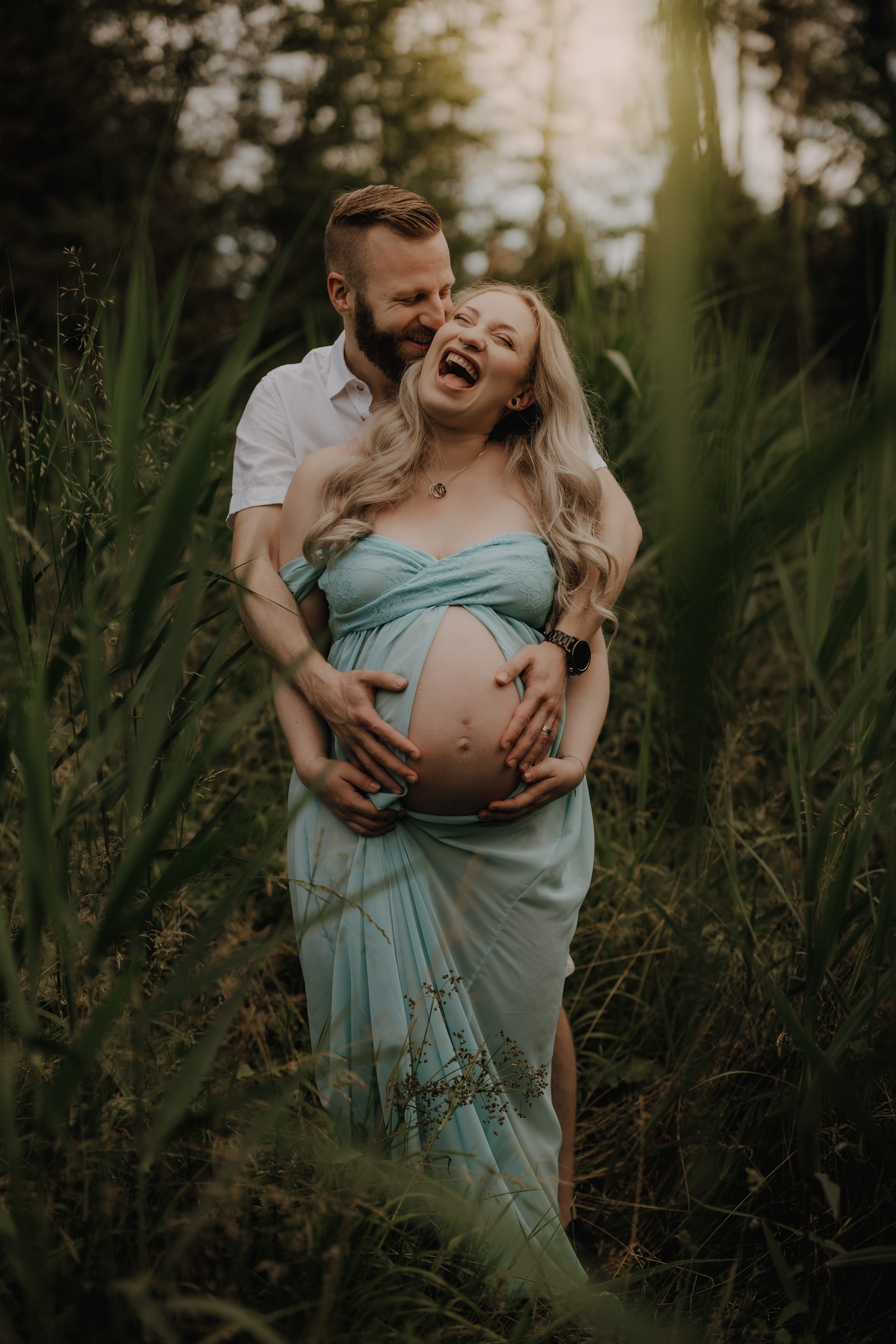 Ein fröhliches Paar steht in einem üppigen, grünen Wald in Baden. Die Frau, die ein hellblaues Kleid trägt, wiegt ihren Babybauch, während der Mann, der ein weißes Hemd trägt, seine Arme von hinten um sie legt. Beide lächeln und lachen während ihres Schwangerschafts-Fotoshootings, umgeben von hohem Gras und Bäumen.