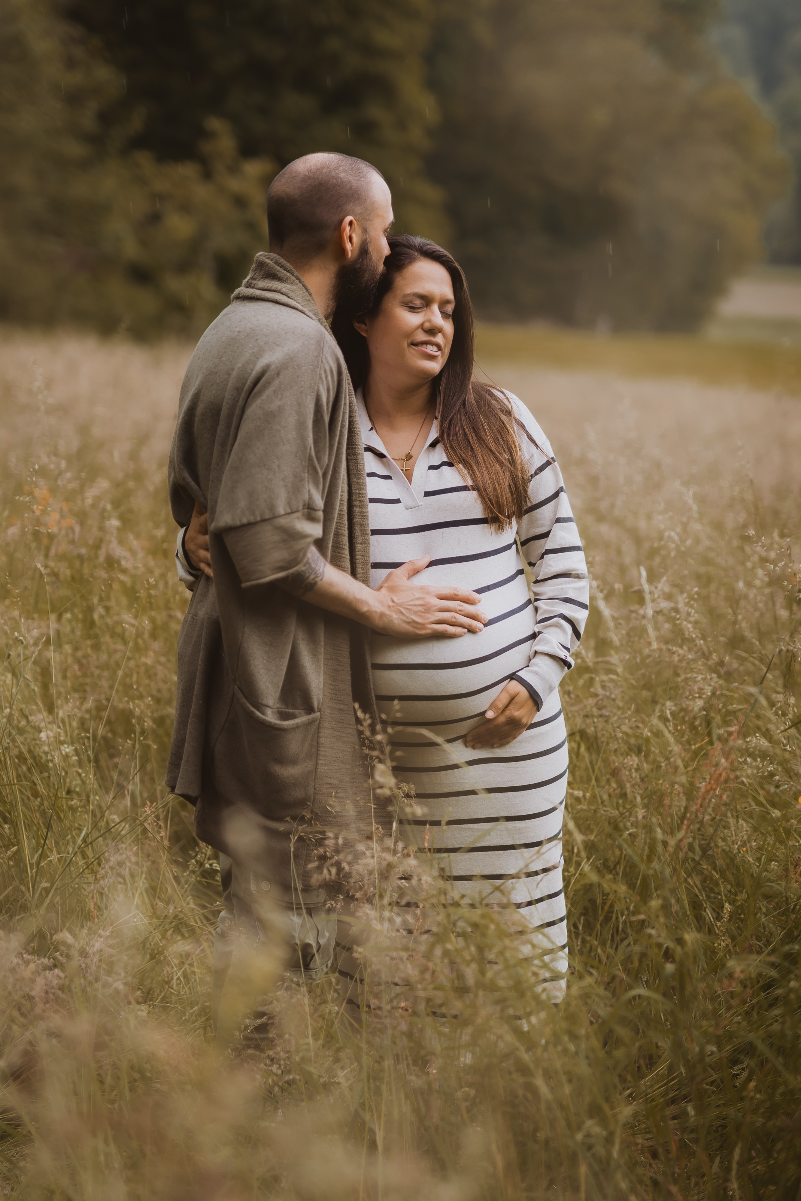 Eine schwangere Frau in einem gestreiften Kleid steht auf einer Wiese und wird von einem Mann von der Seite umarmt. Sie blicken sich liebevoll an, im Hintergrund schaffen Bäume eine ruhige, natürliche Kulisse – ein perfekter Moment, den Tasha Ender während ihres Fotoshootings in Baden eingefangen hat.