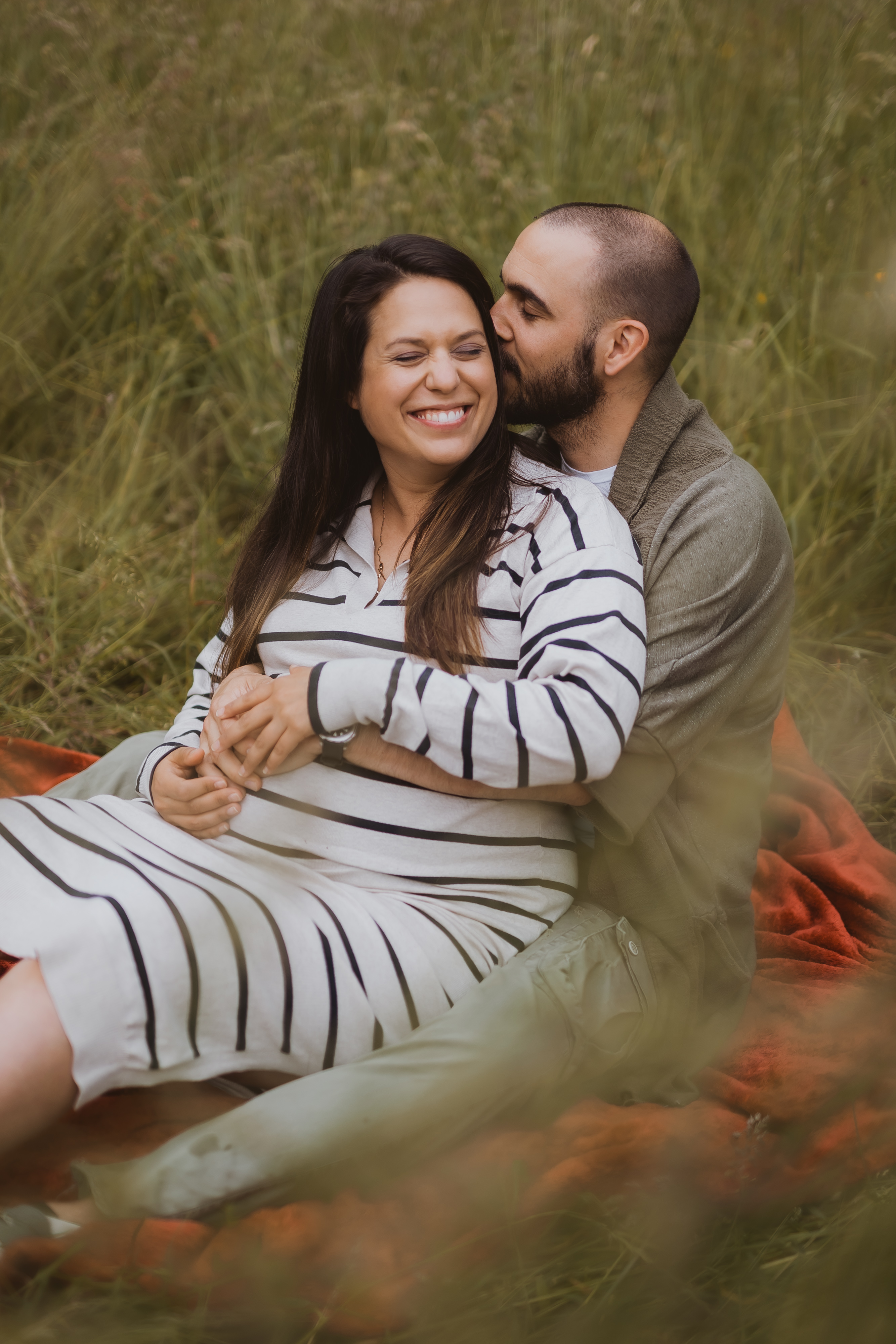 Ein Paar sitzt auf einer orangefarbenen Decke auf einer Wiese. Die Frau, gekleidet in ein weiß-schwarz gestreiftes Kleid, lächelt mit geschlossenen Augen, während der Mann, der einen grünen Pullover trägt, seine Arme um sie legt und sie auf die Wange küsst. Aufgenommen von der Fotografin Tasha Ender, wirken sie glücklich und liebevoll.