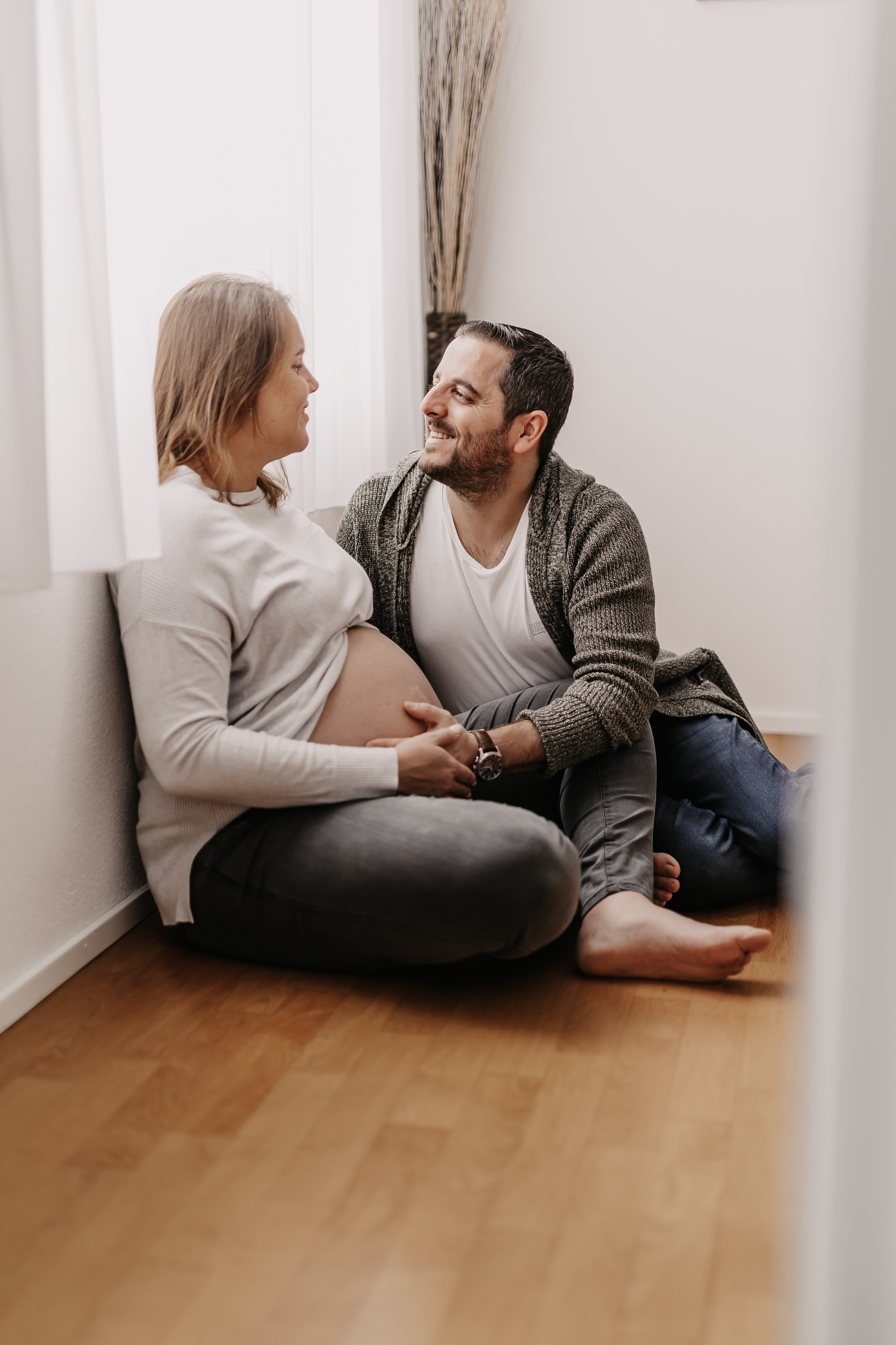 Eine schwangere Frau und ein Mann, beide leger gekleidet, sitzen auf einem Holzboden neben einer weißen Wand und lächeln sich an. Der Mann hält zärtlich die Hand der Frau, während sie ihre Hand auf ihren Babybauch legt und fängt so die intime und freudige Essenz ihres Schwangerschaftsshootings ein.
