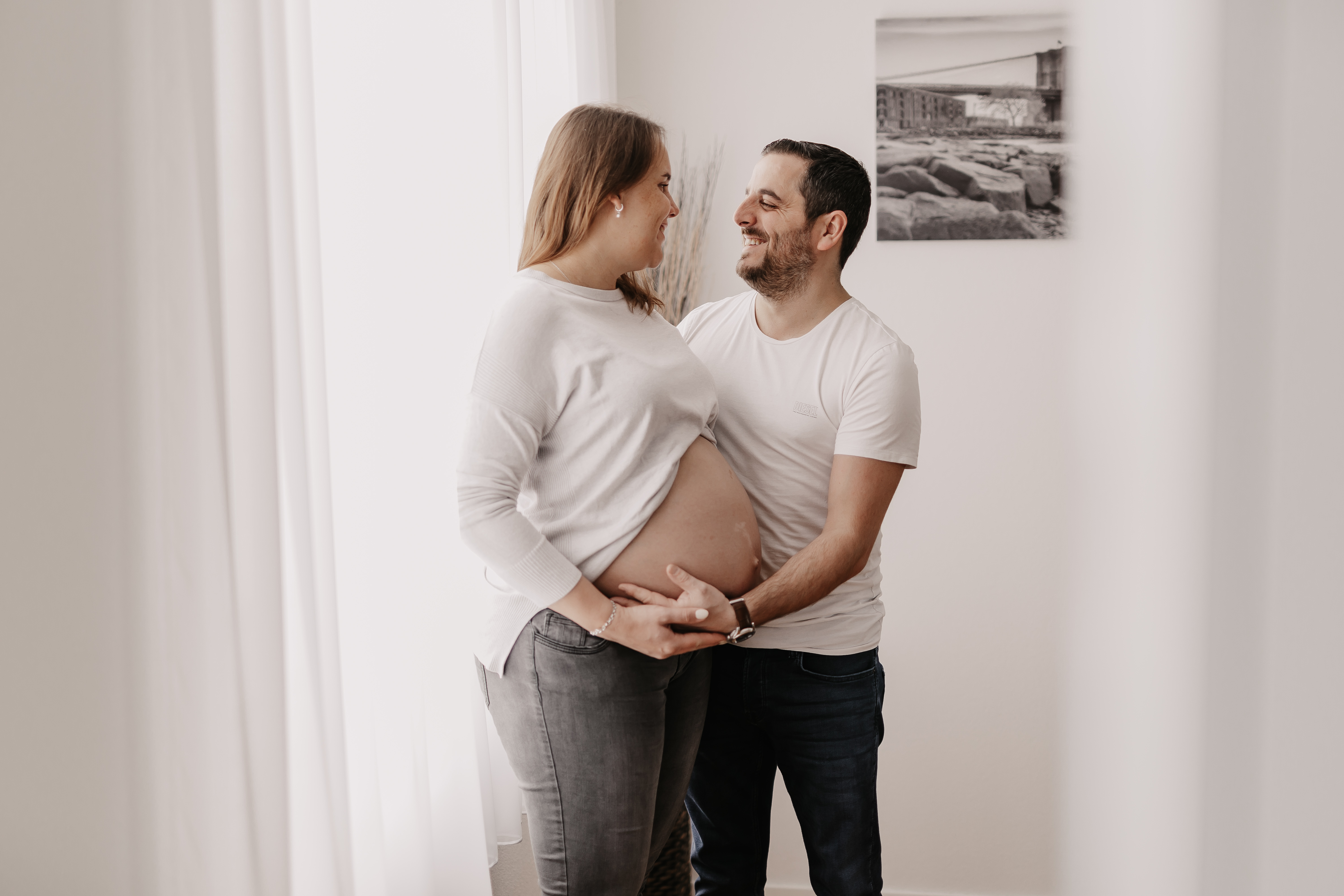 Ein Paar steht in einem hellen Raum, lächelt und schaut sich an. Die Frau, die ihren Babybauch hält, trägt einen hellen Pullover und graue Jeans. Der Mann, der seine Hände auf ihrem Bauch hat, trägt ein weißes Hemd und dunkle Jeans. An der Wand hinter ihnen hängt ein gerahmtes Foto; sie scheinen bereit für ihr Schwangerschaftsshooting.