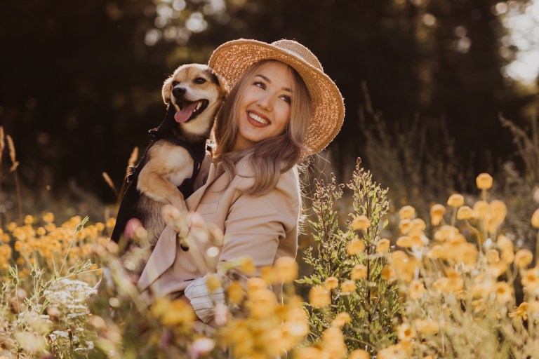Eine Frau mit Strohhut lächelt, während sie einen kleinen Hund auf einer Wiese voller gelber Blumen hält. Das Sonnenlicht fällt durch die Bäume und taucht die Szene in einen warmen Schein. Der Hund sieht glücklich aus und hängt mit seiner heraushängenden Zunge den perfekten Moment fest, den jeder Hundefotograf in Erinnerung behalten wird.