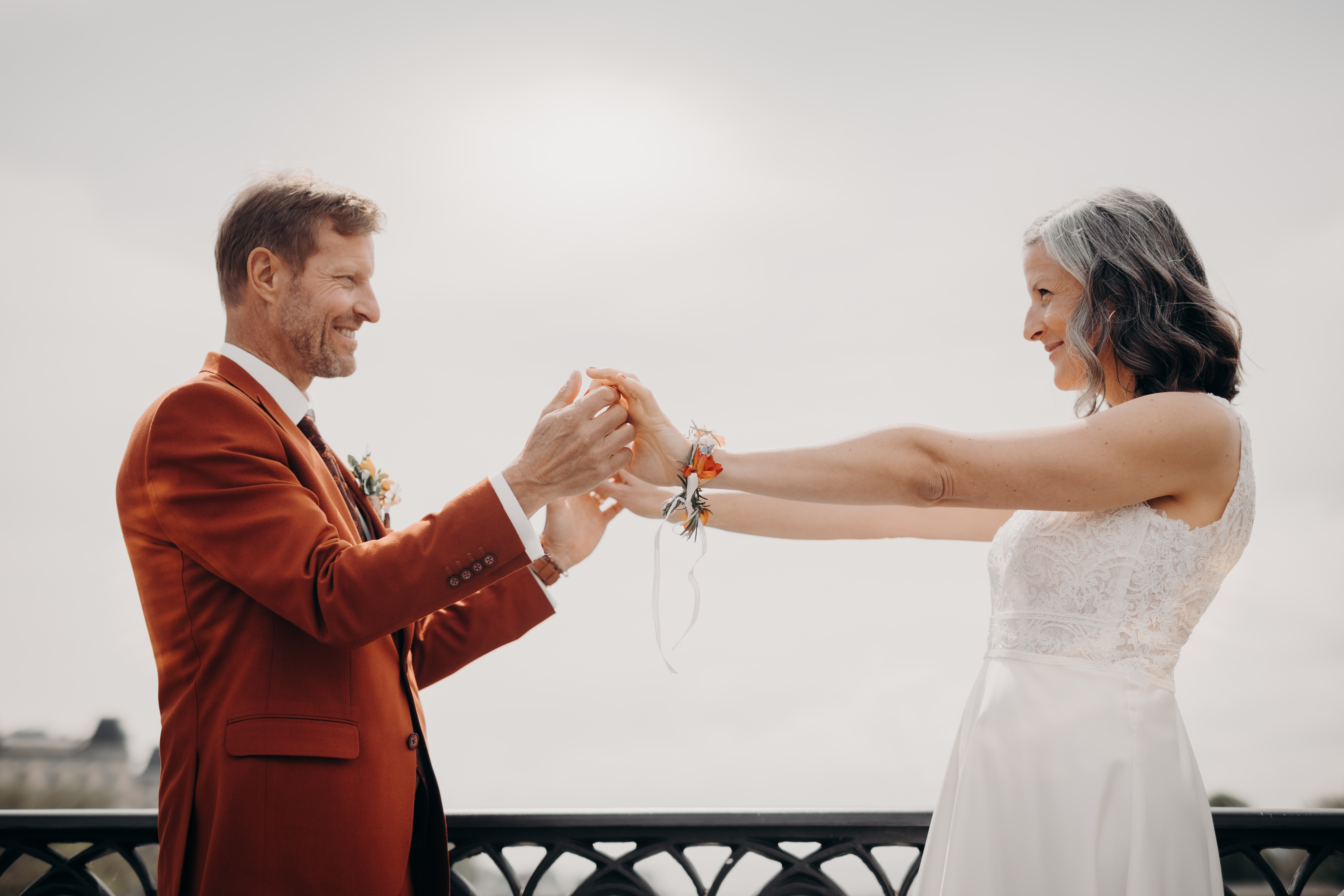 Ein Paar hält sich an den Händen und blickt sich lächelnd an. Der Mann trägt einen orangefarbenen Anzug und die Frau ein weißes Kleid mit ausgestreckten Armen. Beide haben Blumenschmuck an ihren Handgelenken. Sie stehen draußen mit einem weichen, hellen Himmel im Hintergrund – eine perfekte Aufnahme für Hochzeitsfotografie.