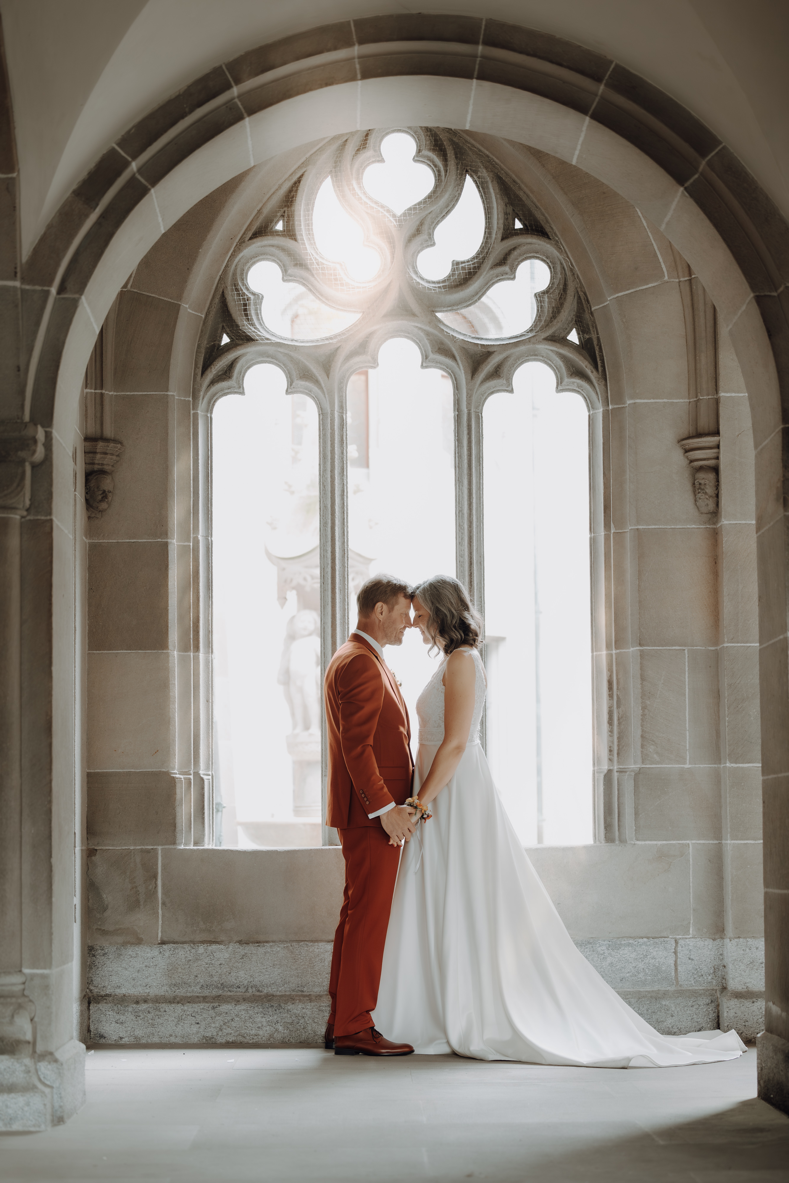 Ein Paar steht einander gegenüber und hält sich an den Händen in einem gewölbten, steingerahmten Fenster mit aufwendigen Mustern. Der Mann trägt einen roten Anzug und die Frau ein weißes Kleid. Sonnenlicht fällt durch das Fenster und beleuchtet sie sanft während ihrer Ziviltrauung in Zürich.