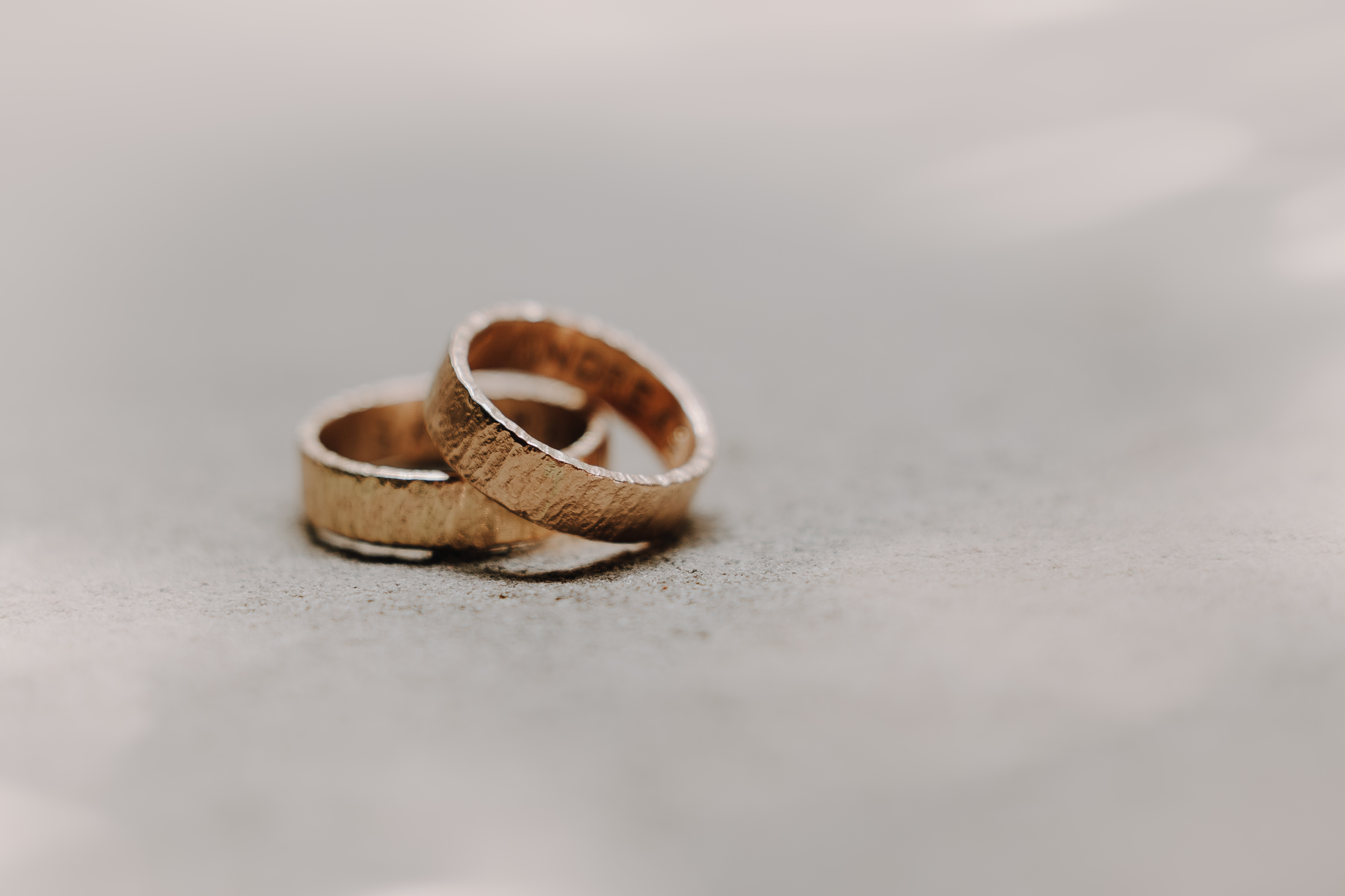 Eine Nahaufnahme von zwei strukturierten goldenen Eheringen, die auf einer glatten, hellen Oberfläche liegen. Beide Ringe haben eine leicht gebürstete Oberfläche und die sanfte Beleuchtung erzeugt eine warme, intime Atmosphäre. Die Ringe sind so positioniert, dass sie sich leicht überlappen, was die Essenz von Tasha Enders Züricher Ziviltrauung einfängt.