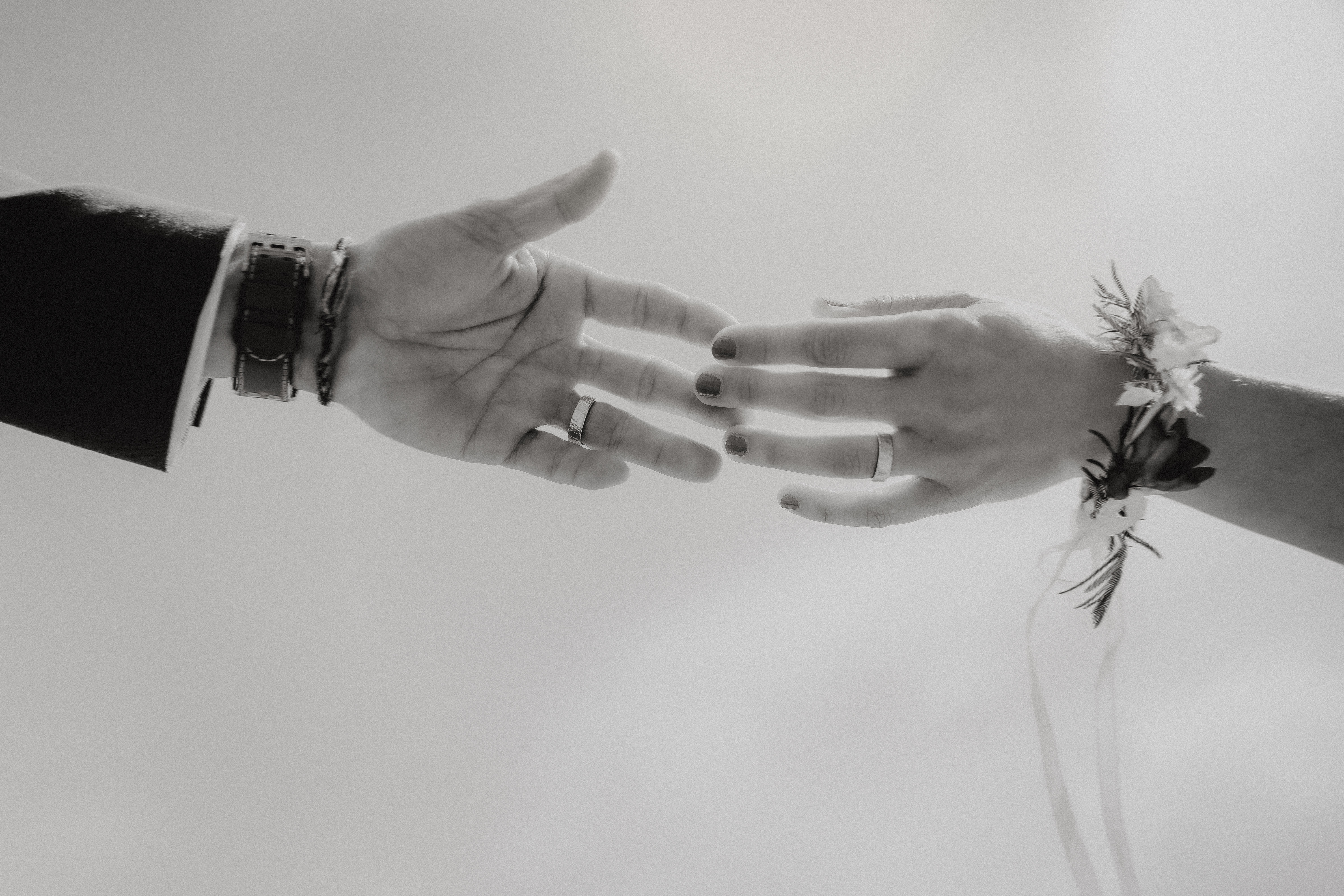 Nahaufnahme in Schwarzweiß von zwei Händen, die sich einander nähern. Die linke Hand ist männlich und trägt eine Uhr und Ringe, während die rechte Hand weiblich ist und mit einem Blumenanstecker und lackierten Nägeln geschmückt ist. Diese atemberaubende Aufnahme einer Hochzeitsfotografin fängt den intimen Moment perfekt vor einem sanft verschwommenen Hintergrund ein.