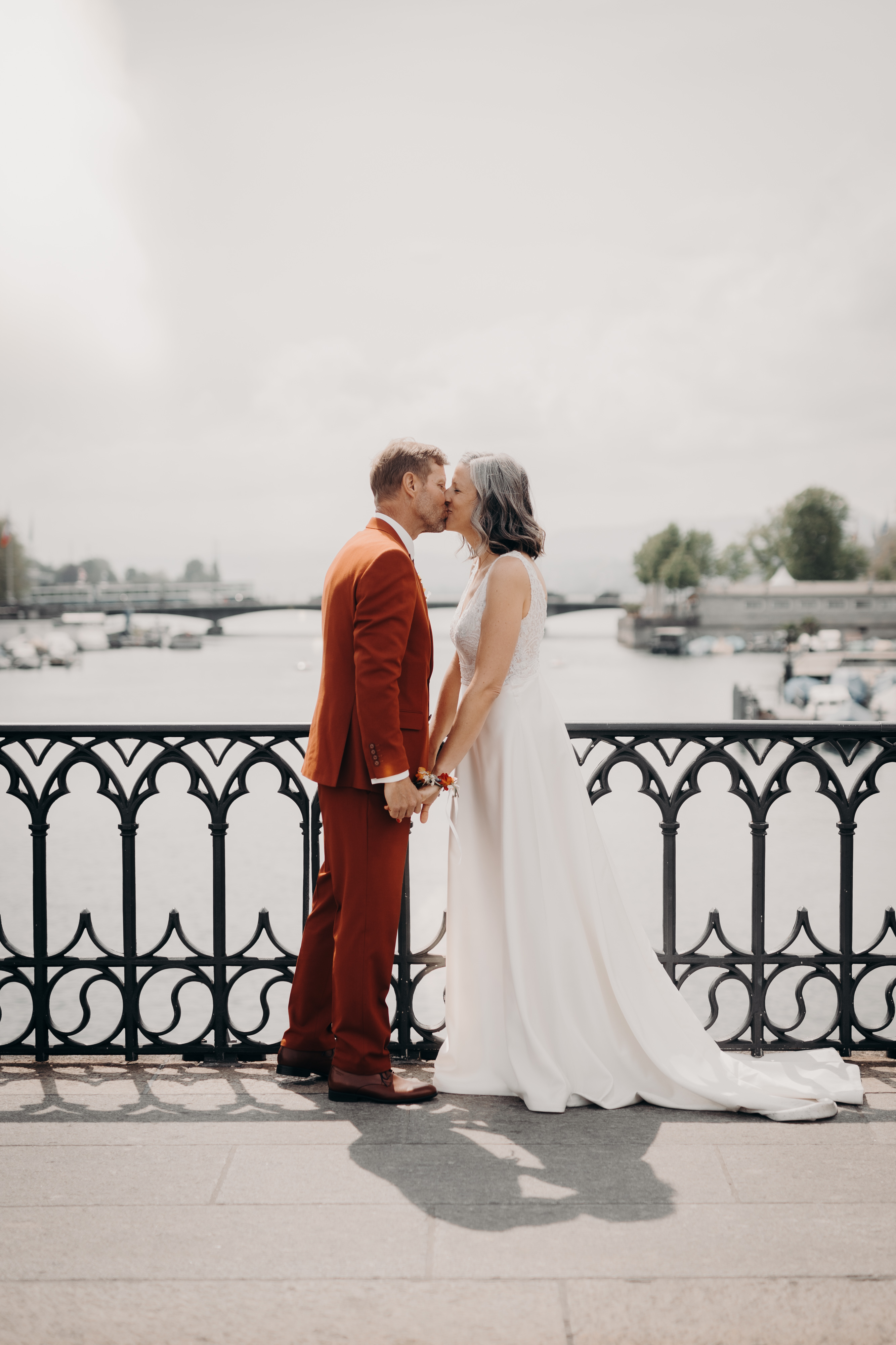 Eine Braut in einem weißen Kleid und ein Bräutigam in einem rotbraunen Anzug küssen sich und halten sich an den Händen. Sie stehen auf einer Brücke mit einem verzierten Geländer, im Hintergrund sieht man Zürichs malerische Uferpromenade und Boote. Der Himmel ist bedeckt und fängt die Essenz der Hochzeitsfotografie während ihrer Ziviltrauung ein.