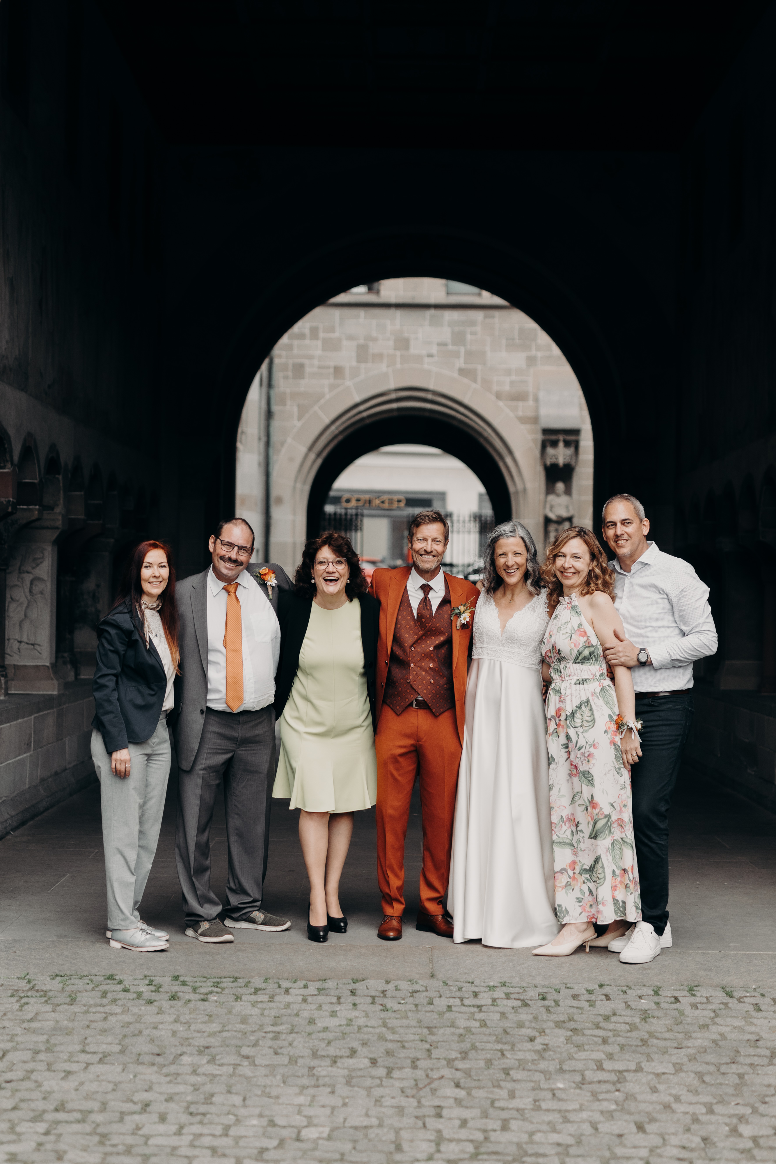 Eine Gruppe von sieben Personen, formell gekleidet, posiert gemeinsam im Freien. Die Personen lächeln, und ein Paar in der Mitte scheint im Mittelpunkt zu stehen, wobei der Mann einen rotbraunen Anzug und die Frau ein weißes Kleid trägt. Ein Torbogen und ein Steingebäude bieten eine bezaubernde Kulisse für diese Hochzeit in Zürich.