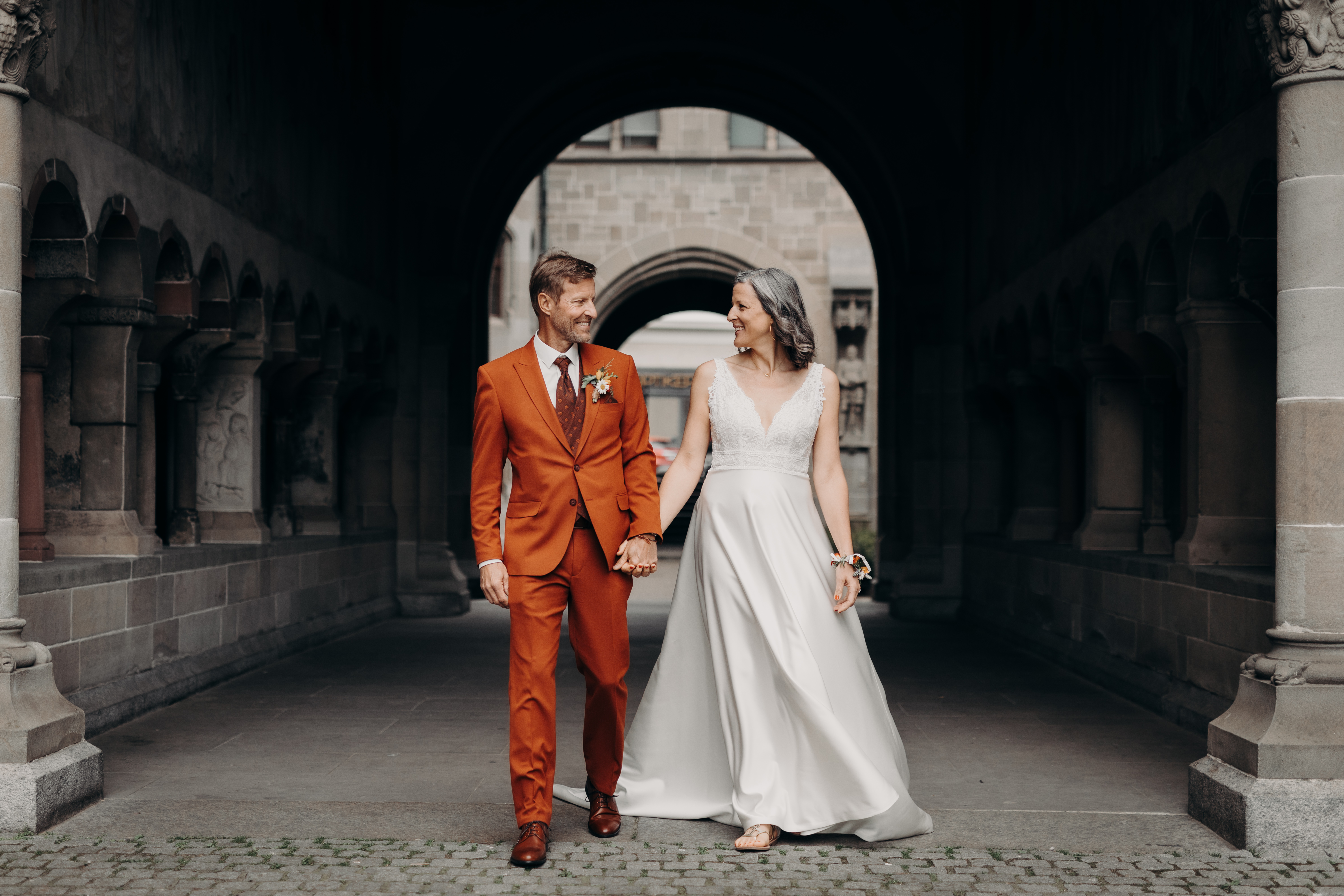 Braut und Bräutigam halten Händchen und lächeln sich an, während sie durch einen steinernen Torbogen gehen. Der Bräutigam trägt einen rostfarbenen Anzug und die Braut ein weißes ärmelloses Kleid. Die Kulisse ihrer Hochzeit ist ein historisches Gebäude mit komplexer Architektur, wunderschön eingefangen von Tasha Ender.