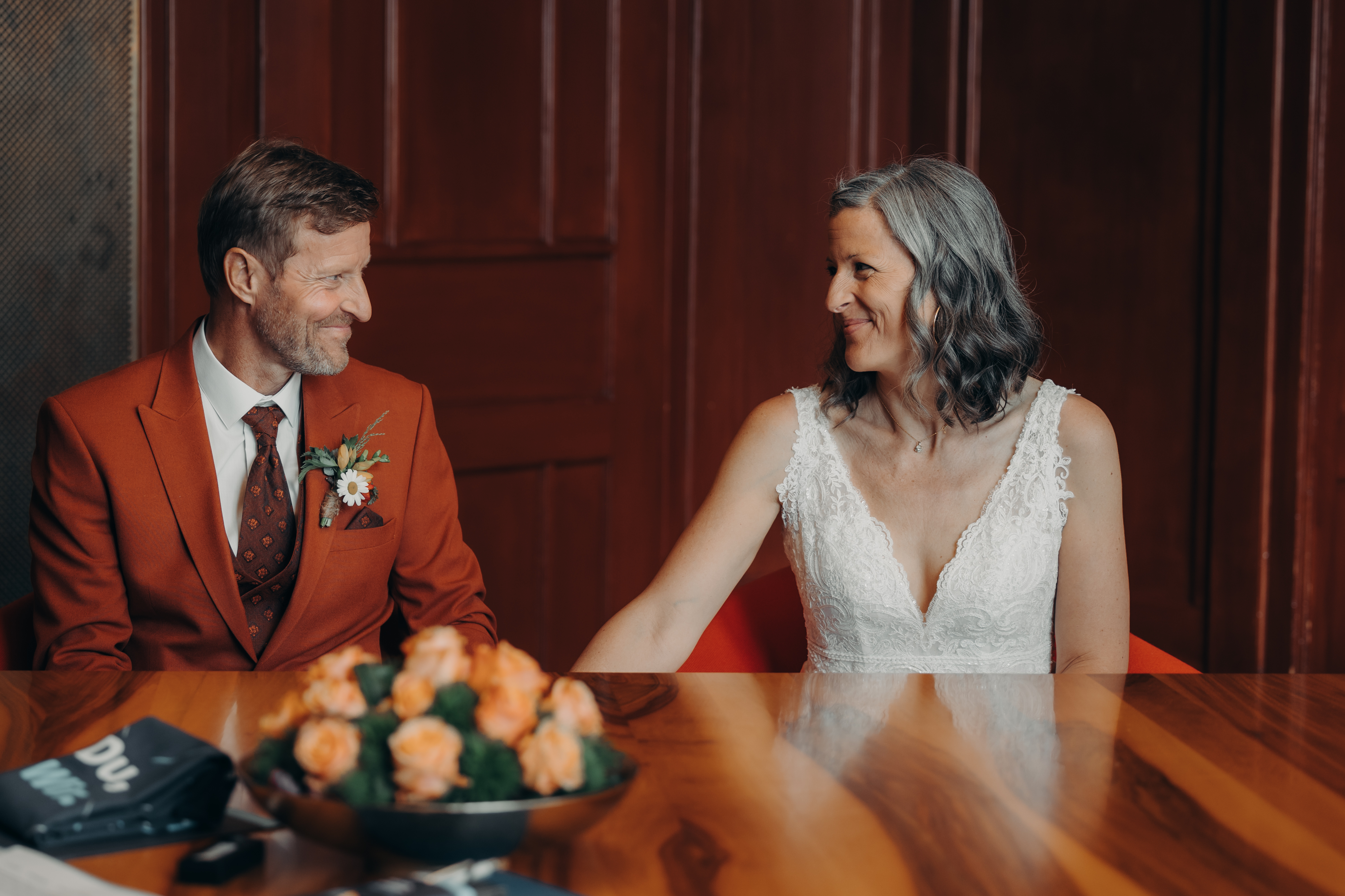 Ein fröhliches Paar sitzt zusammen an einem Holztisch in Zürich und lächelt sich herzlich an. Der Mann trägt einen rostfarbenen Anzug und eine gemusterte Krawatte, während die Frau in einem weißen Spitzenkleid glänzt. Ein Strauß orangefarbener Blumen, aufgenommen von Tasha Ender Photography für ihre Zivilhochzeit, schmückt den Tisch im Vordergrund.