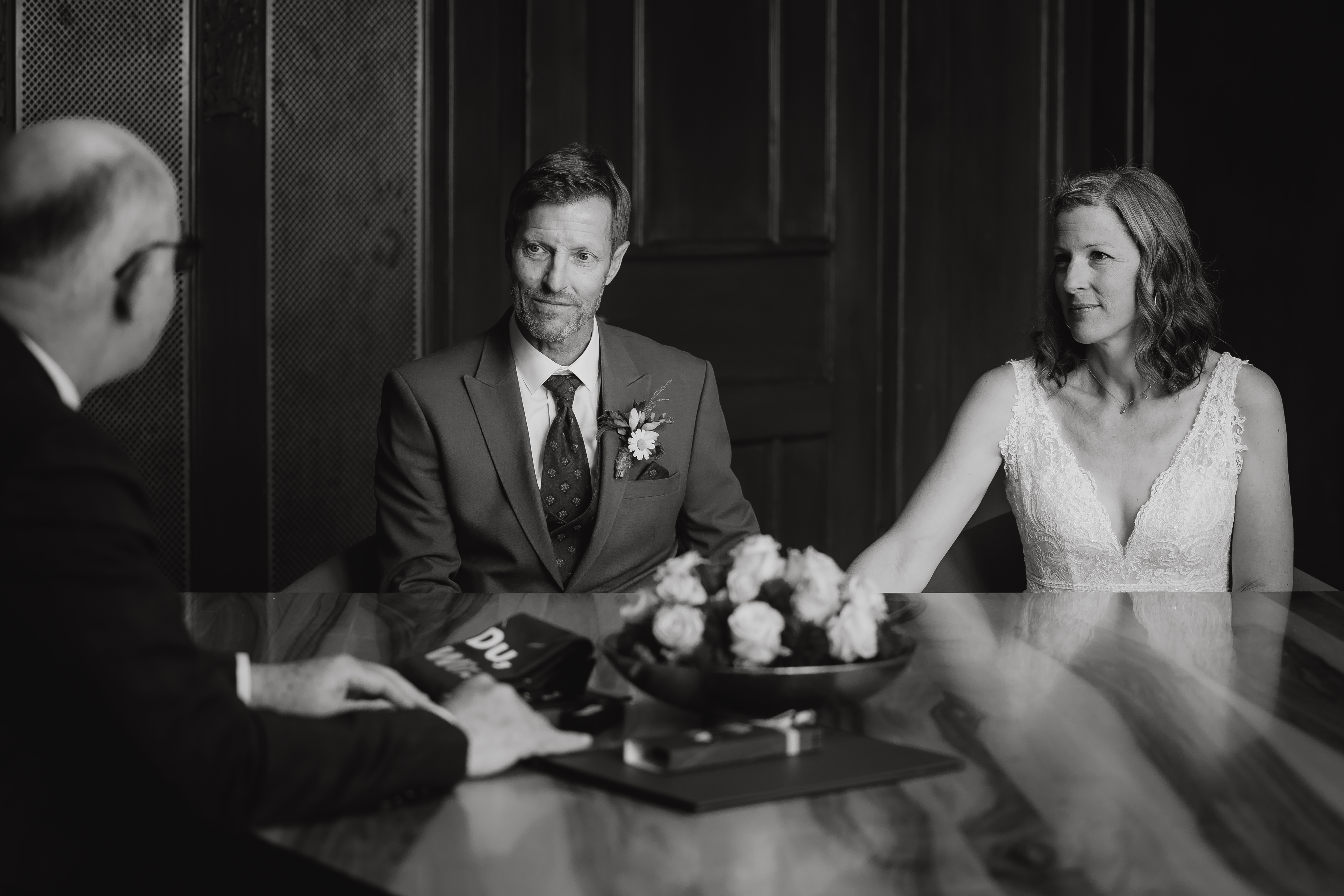 Ein Paar in Hochzeitskleidung, der Bräutigam im Anzug und die Braut im weißen Kleid, sitzt an einem polierten Holztisch und hält sich an den Händen. Ein Mann im Anzug, möglicherweise der Geistliche, ist von hinten über den Tisch zu sehen. Ein Blumenstrauß steht während ihrer Ziviltrauung in Zürich in der Mitte des Tisches. Der Moment wurde wunderschön von Hochzeitsfotografie von Tasha Ender Photography eingefangen.