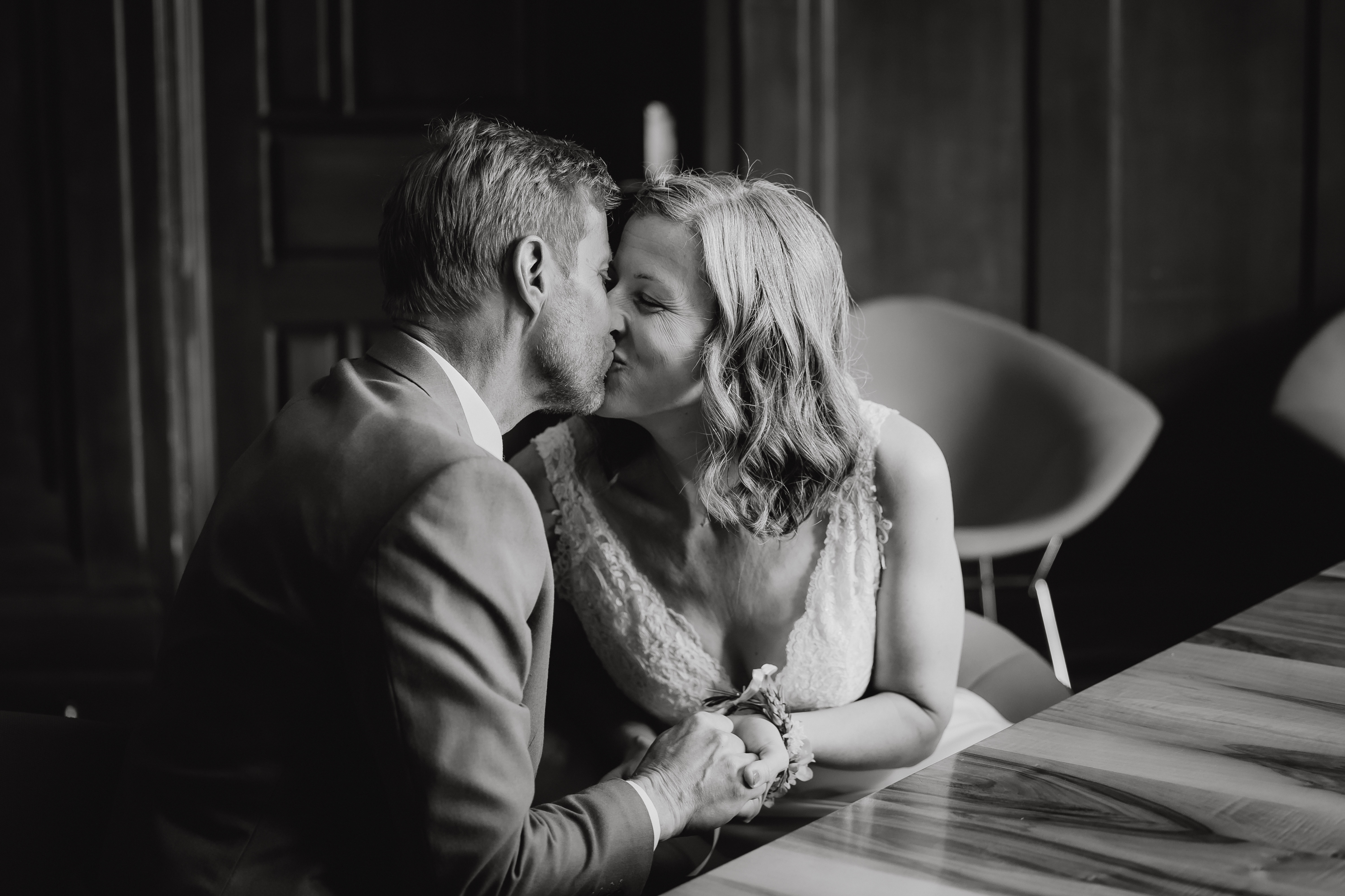 Ein Paar in Hochzeitskleidung küsst sich, während es sich über einen Holztisch hinweg an den Händen hält. Das Schwarz-Weiß-Foto von Tasha Ender Photography's Hochzeitsfotografie fängt einen intimen und freudigen Moment während ihrer Zivilhochzeit in Zürich ein. Der Hintergrund ist ein schwach beleuchteter Raum mit Holzpaneelen und modernen Stühlen.