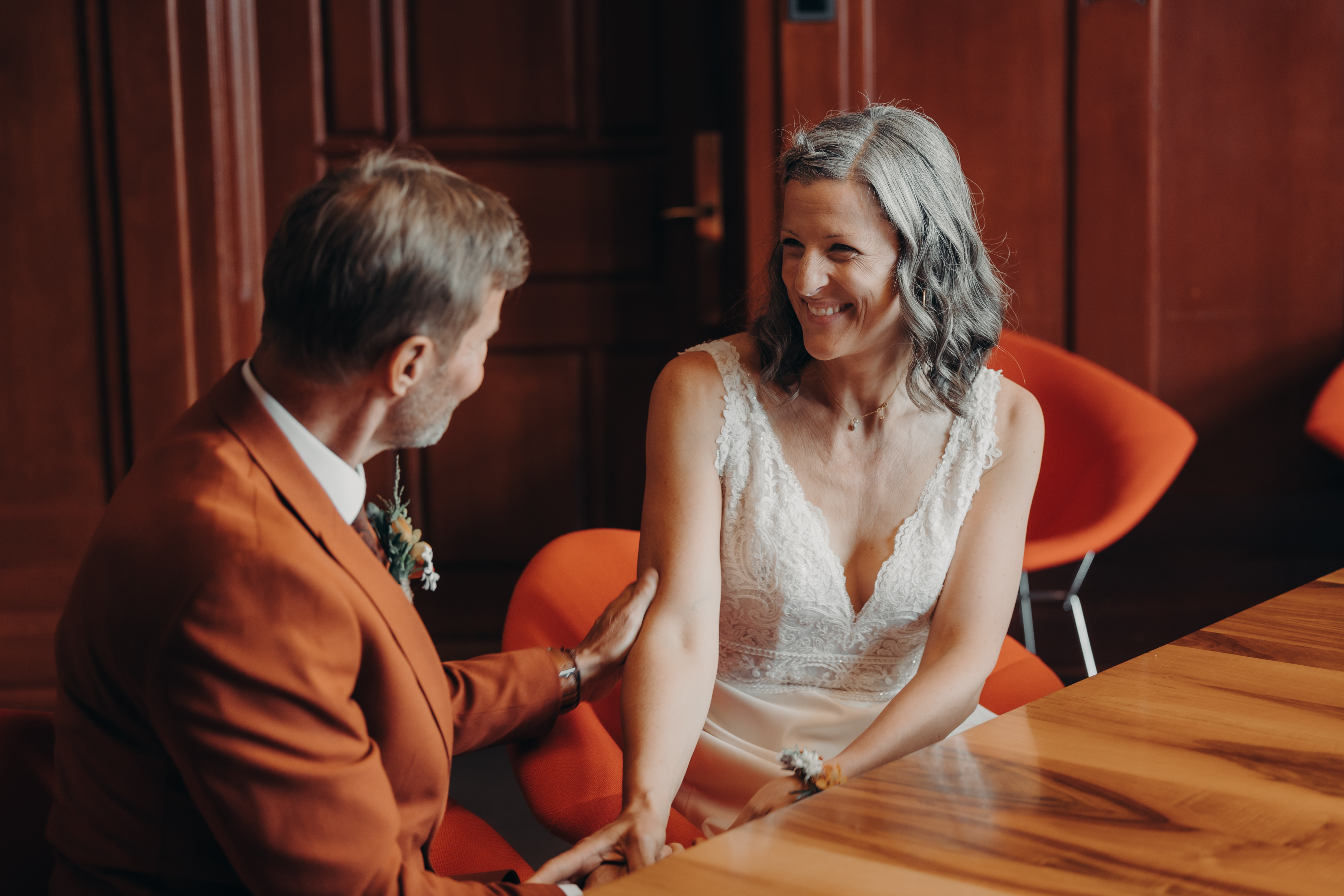 Eine lächelnde Braut mit grauem Haar in einem weißen Spitzenkleid sitzt an einem Holztisch, während ein bärtiger Bräutigam in einem orangefarbenen Jackett sanft ihren Arm hält. Sie schauen sich während ihrer Zivilhochzeit in Zürich liebevoll an, wobei der warm beleuchtete Raum die rot-orangen Stühle im Hintergrund hervorhebt. Dieser wunderschöne Moment wurde von Tasha Ender Photography's Hochzeitsfotografie festgehalten.