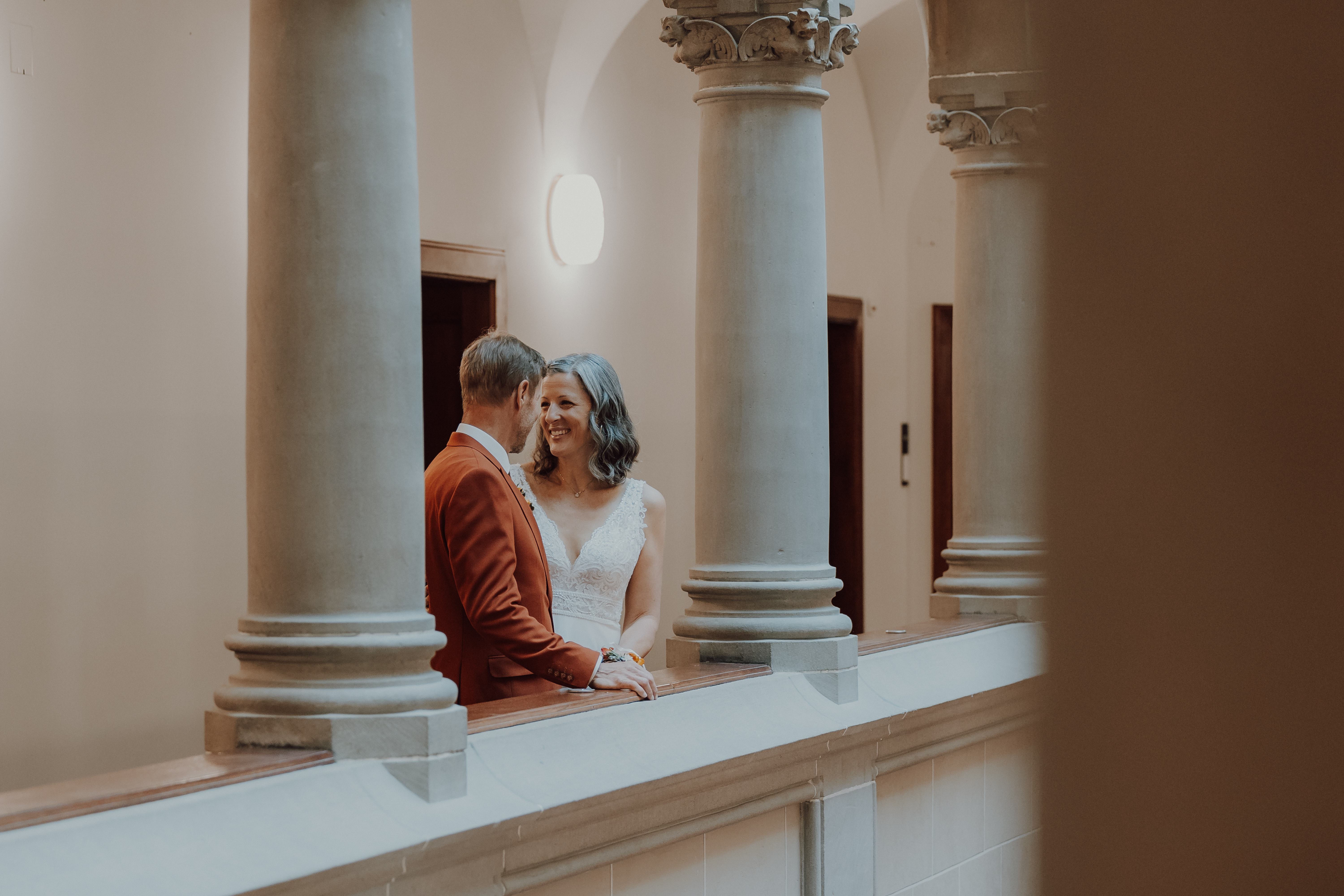 Hier ist der überarbeitete Satz unter Verwendung der angegebenen Produktdaten:
Ein Paar lächelt sich auf einem Innenbalkon mit Säulen an; der Mann trägt eine rote Jacke und die Frau ein ärmelloses weißes Kleid. Sanftes Licht schafft eine warme Atmosphäre, perfekt für ihre Ziviltrauung, wunderschön eingefangen von Tasha Ender Photography's Hochzeitsfotografie.