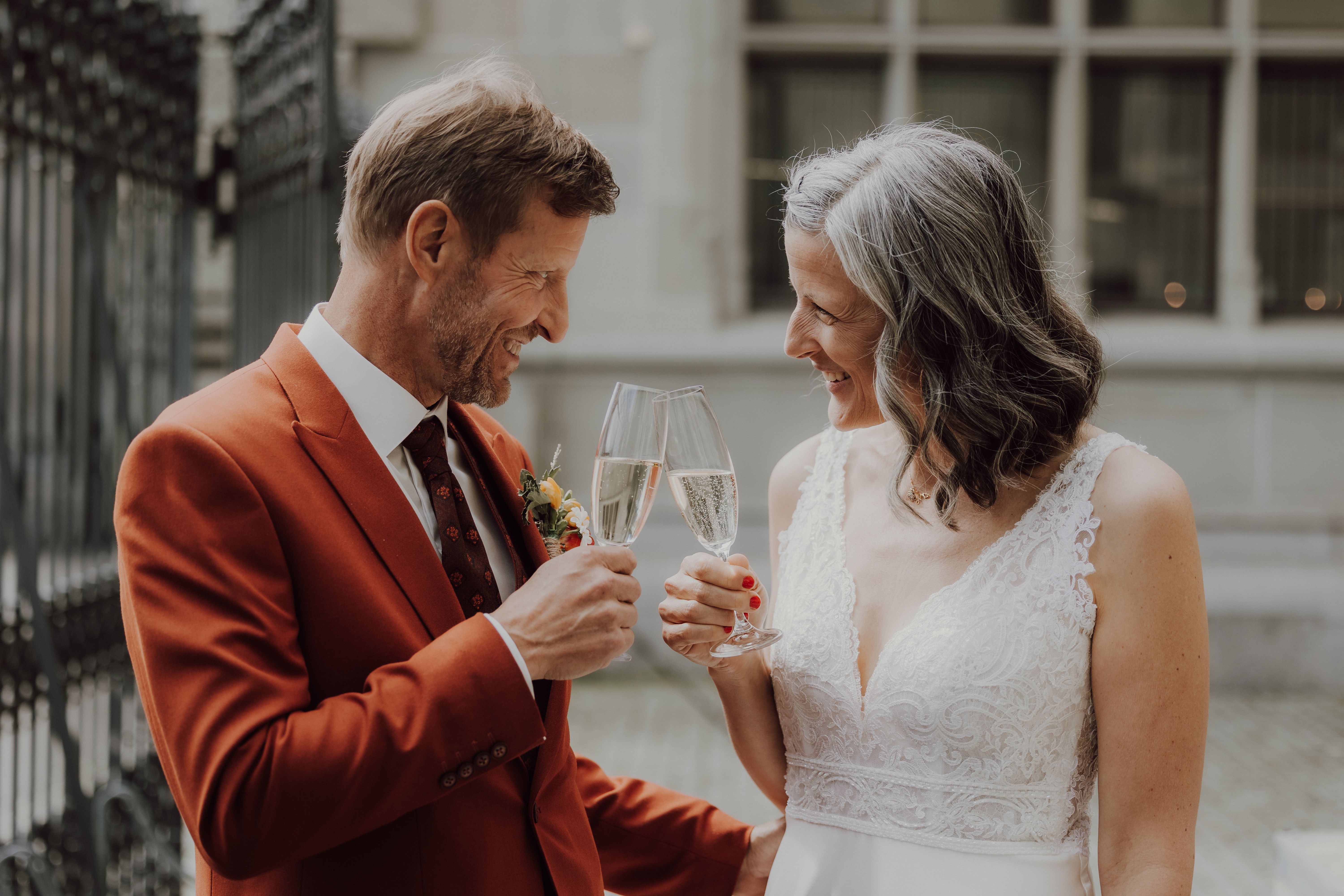 Ein Paar feiert im Freien in Zürich, lächelt und stößt mit Champagnergläsern an. Der Mann, elegant gekleidet in einem roten Anzug, und die Frau, strahlend in einem weißen Spitzenhochzeitskleid, teilen einen freudigen Moment. Sie stehen nach ihrer Ziviltrauung vor einem Gebäude mit großen Fenstern und Eisentoren. Dieser wunderschöne Moment wurde vom Hochzeitsfotografie-Service von Tasha Ender Photography fachmännisch festgehalten.