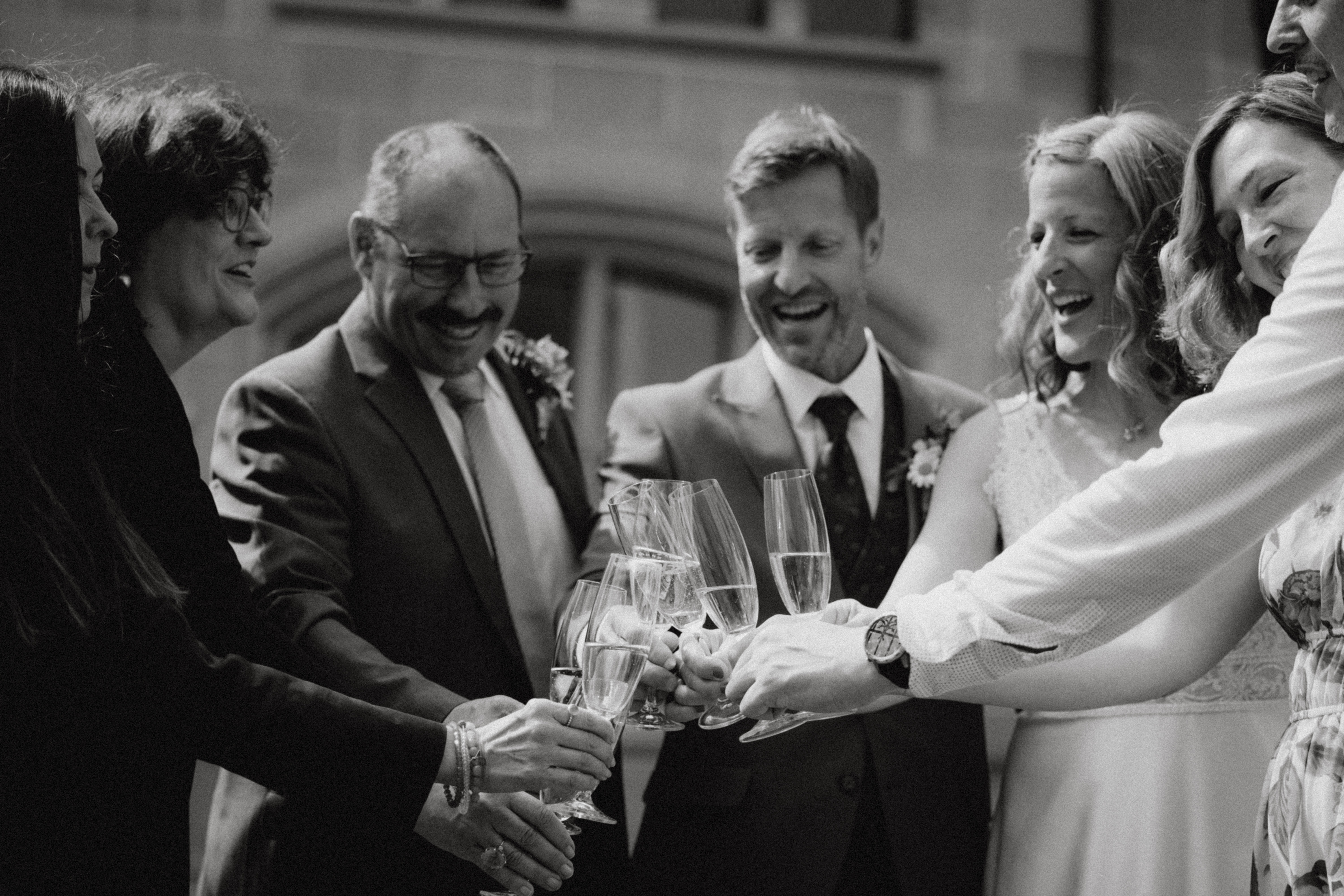 Eine Gruppe von Menschen in formeller Kleidung steht lächelnd und lachend im Kreis und erhebt ihre Champagnergläser zum Anstoßen. Der Moment, der während einer Ziviltrauung in Zürich von Tasha Ender Photography's Hochzeitsfotografie festgehalten wurde, ist in Schwarzweiß gehalten und verleiht der feierlichen Szene ein zeitloses Gefühl.