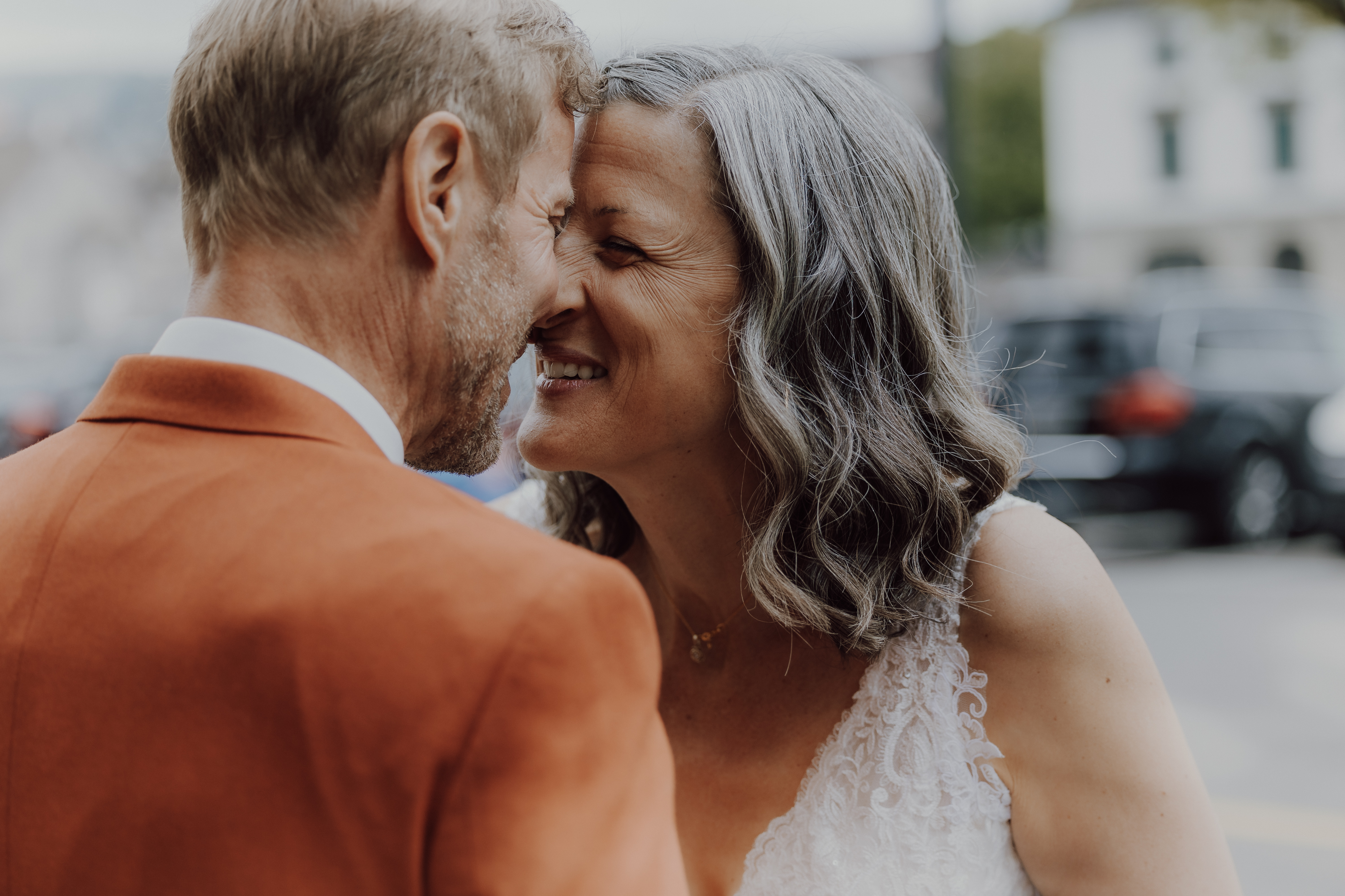 Ein älteres Paar in formeller Kleidung teilt einen zärtlichen Moment während seiner Zivilhochzeit, lächelt und lehnt sich eng aneinander. Die Frau trägt ein weißes Spitzenkleid und der Mann ein orangefarbenes Jackett. Der Hintergrund ist leicht verschwommen und deutet auf eine Szene im Freien in Zürich hin – alles wunderschön eingefangen vom Hochzeitsfotografie-Service von Tasha Ender Photography.