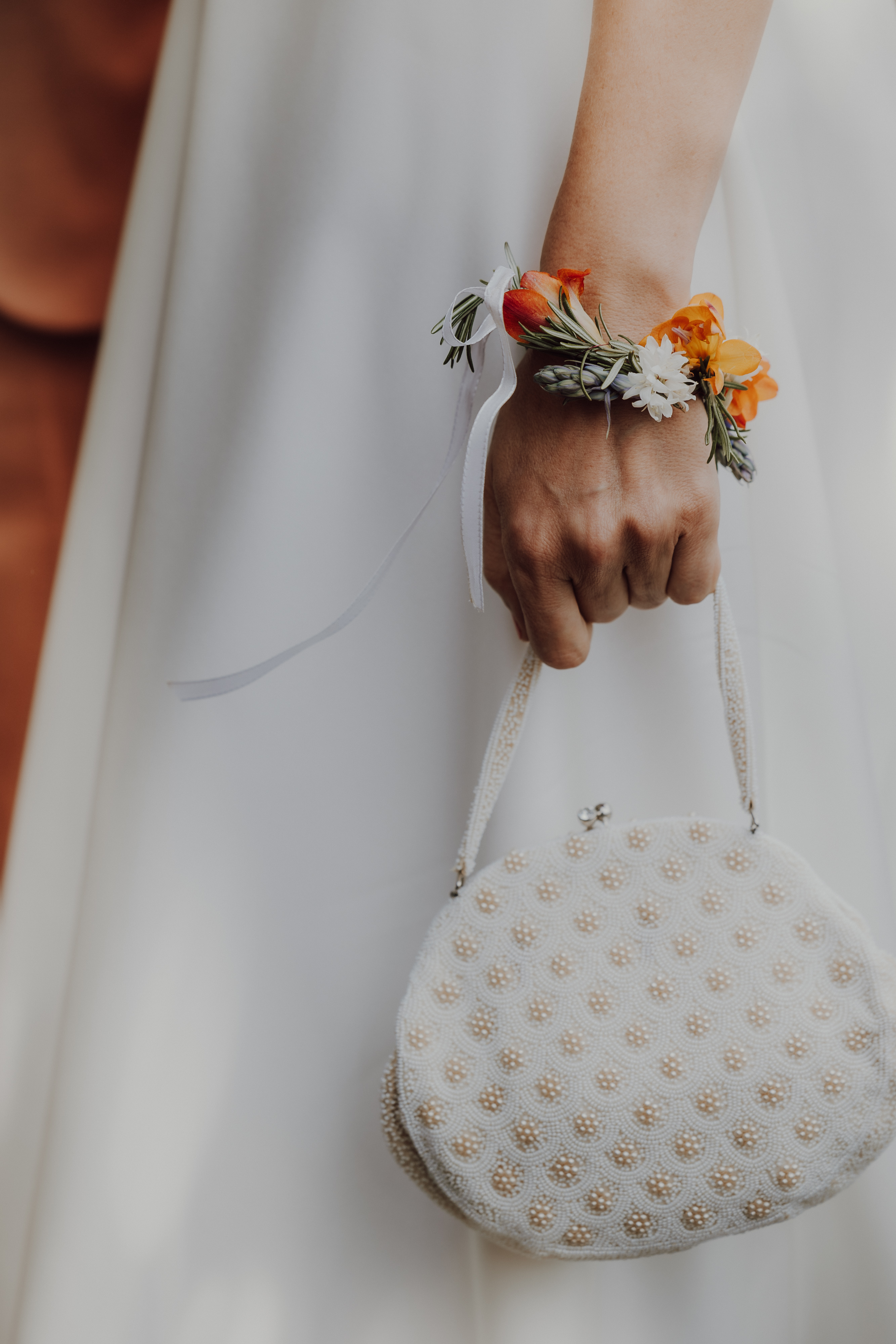 Eine Nahaufnahme von Tasha Ender Photography zeigt eine Person, die während einer Hochzeitsfotografie-Sitzung für eine Zivilhochzeit in Zürich eine kleine, weiße Handtasche mit Perlen in der rechten Hand hält. Sie trägt ein weißes Kleid und ihr Handgelenk ist mit einem farbenfrohen Blumenarmband mit orangefarbenen, weißen und gelben Blumen vor einem sanft verschwommenen Hintergrund geschmückt.