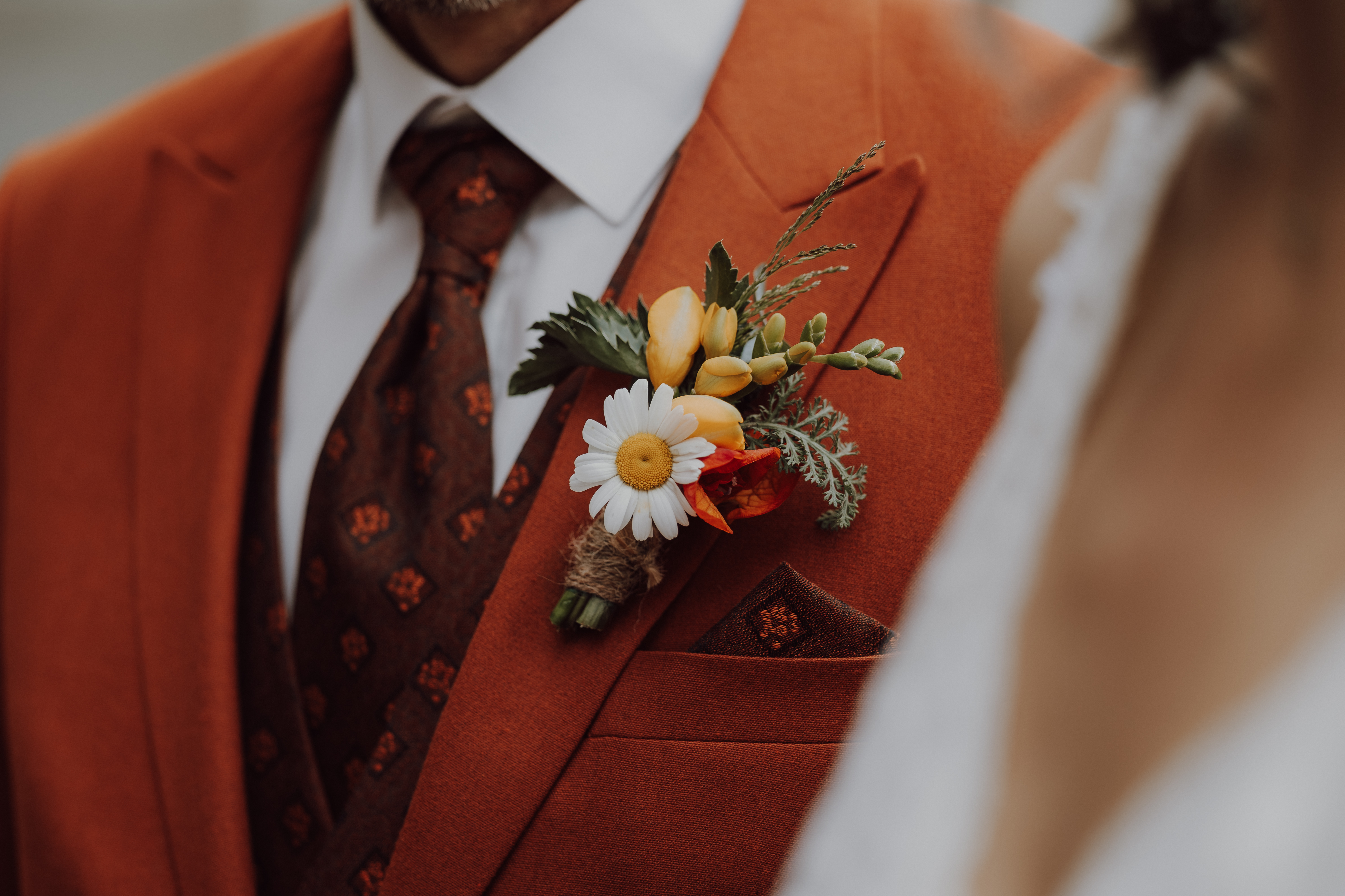 Eine Nahaufnahme von Tasha Ender Photography's Hochzeitsfotografie zeigt eine Person in einem orangefarbenen Anzug und einer gemusterten Krawatte bei einer Zivilhochzeit in Zürich. Die farbenfrohe Ansteckblume, geschmückt mit einem weißen Gänseblümchen, gelben Knospen und roten Akzenten, sticht deutlich hervor. Im rechten Hintergrund ist eine weitere Person teilweise sichtbar und leicht verschwommen.