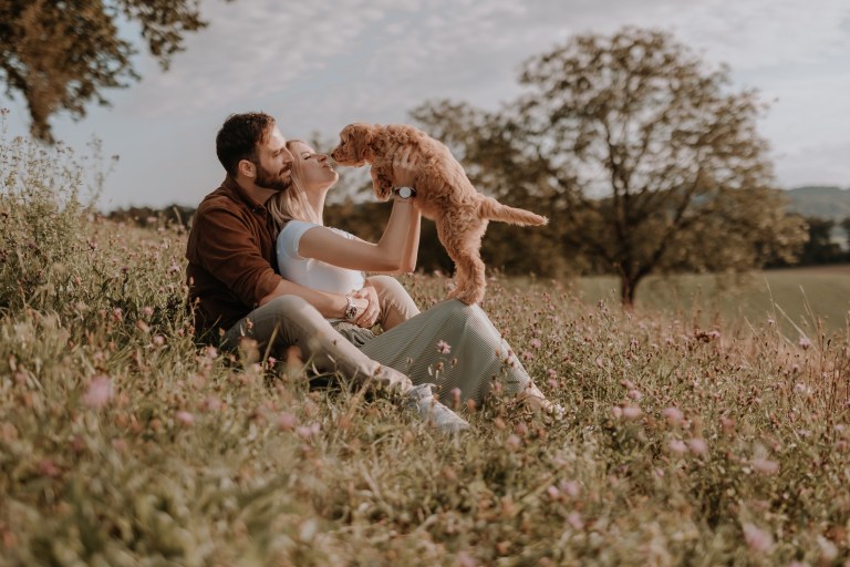Ein Paar sitzt auf einem grasbewachsenen Hügel mit Wildblumen und lächelt, während es mit einem kleinen braunen Hund spielt. Die Frau hält den Hund zu ihrem Gesicht hoch, während der Mann sie von hinten umarmt. Im Hintergrund sind Bäume und ein offenes Feld unter einem teilweise bewölkten Himmel zu sehen, perfekt für ein Welpenshooting mit einem talentierten Hundefotografen.
