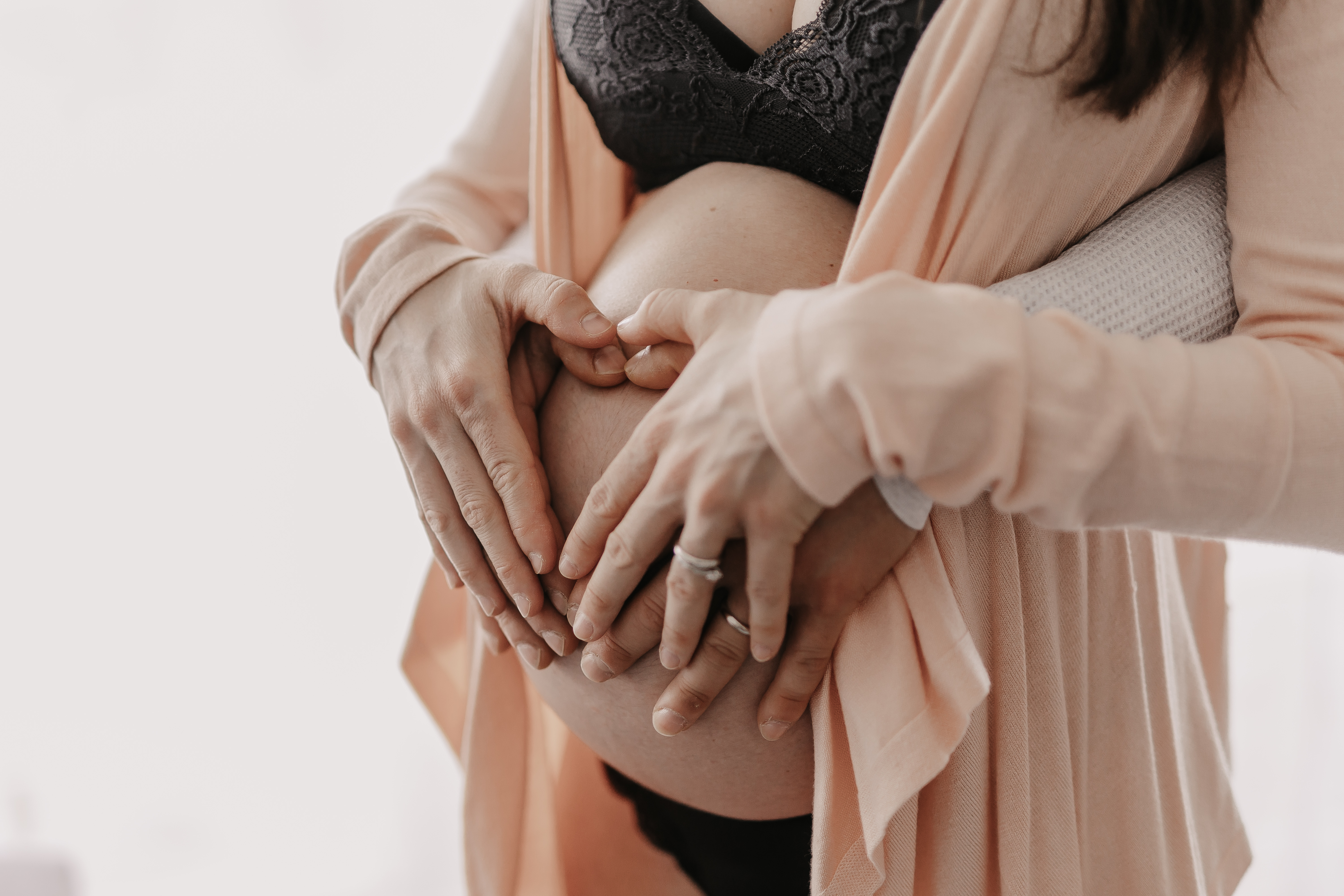 Eine Nahaufnahme einer schwangeren Frau in einem pfirsichfarbenen Morgenmantel, deren Hände auf ihrem nackten Bauch ruhen und mit ihren Fingern ein Herz bilden. Eine andere Person steht hinter ihr und legt ihre Hände ebenfalls auf ihren Bauch, sodass ein größeres Herz entsteht – ein perfekter Moment, eingefangen während dieses Babybauch-Fotoshootings.
