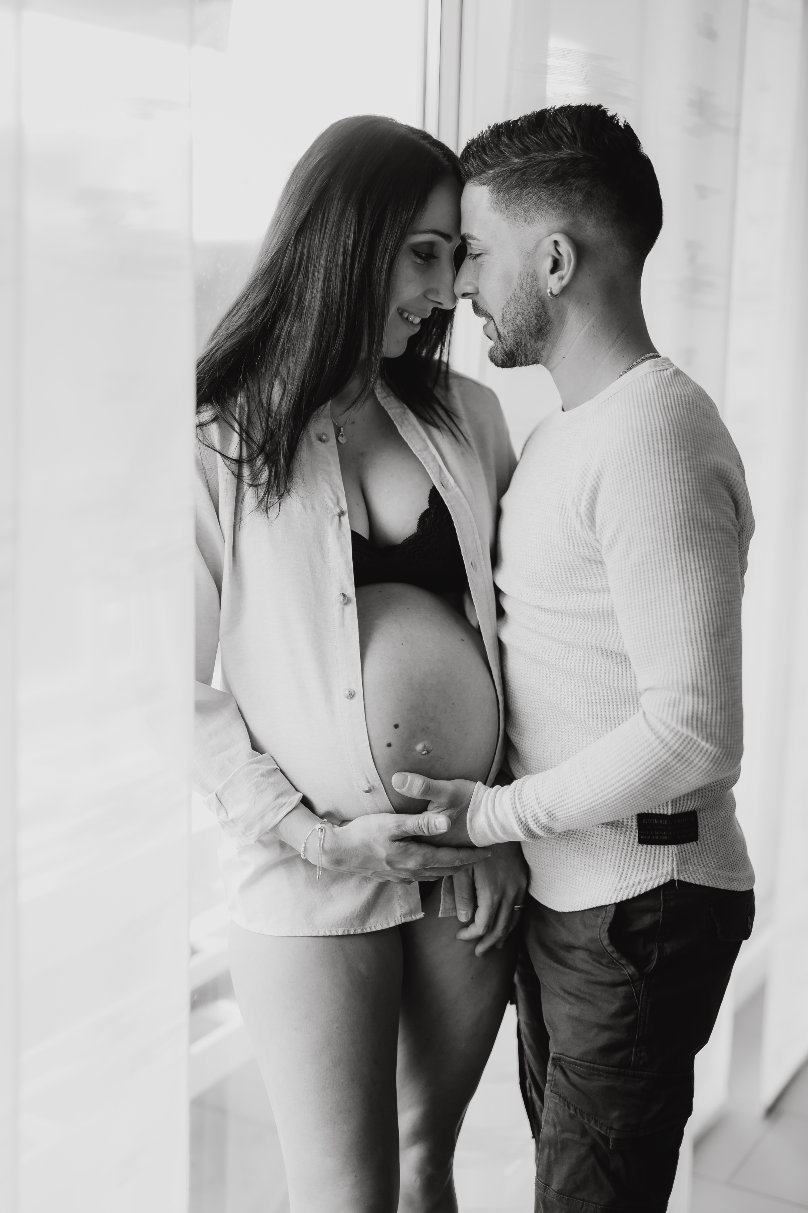 Eine schwangere Frau und ein Mann stehen während eines Babybauch-Fotoshootings an einem Fenster. Das Hemd der Frau ist aufgeknöpft und zeigt ihren Babybauch, den der Mann sanft berührt. Sie blicken sich liebevoll an und ihre Stirnen berühren sich in dieser atemberaubenden Schwarz-Weiß-Aufnahme fast.