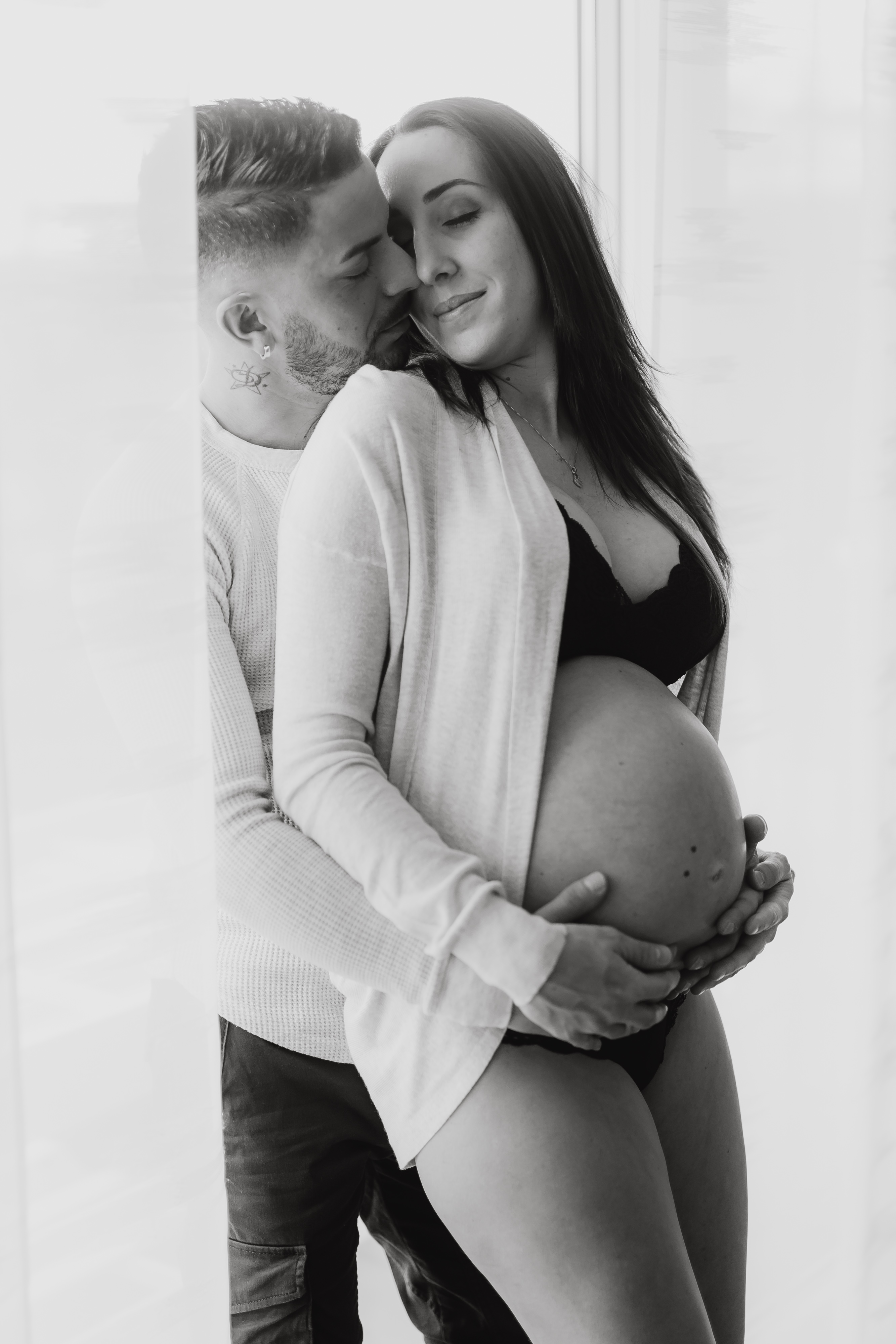 Ein Schwarzweißfoto eines Paares während eines Schwangerschafts-Fotoshootings. Der Mann steht hinter der schwangeren Frau und küsst sie liebevoll auf die Wange. Die Frau, die einen BH und eine aufgeknöpfte Strickjacke trägt, wiegt ihren Babybauch. Beide sehen zufrieden aus und teilen einen intimen Moment an einem Fenster.
