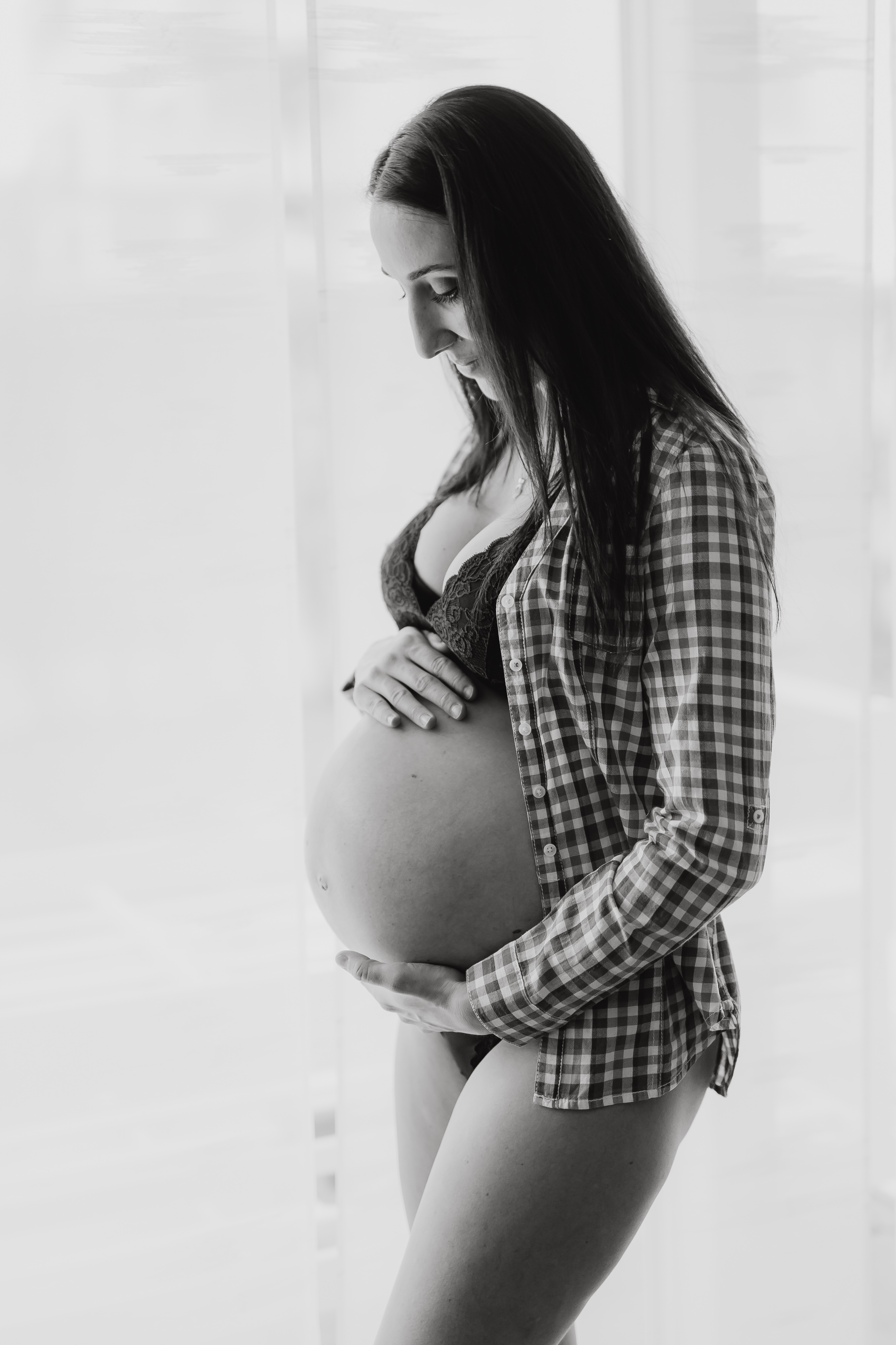 Eine schwangere Frau steht vor einem Fenster und hält ihren Babybauch mit beiden Händen. Sie trägt ein kariertes Hemd und schwarze Dessous. Das Schwarzweißfoto fängt ihren friedlichen Gesichtsausdruck und das natürliche Licht ein, das beim Babybauch-Fotoshooting durch die Vorhänge fällt.