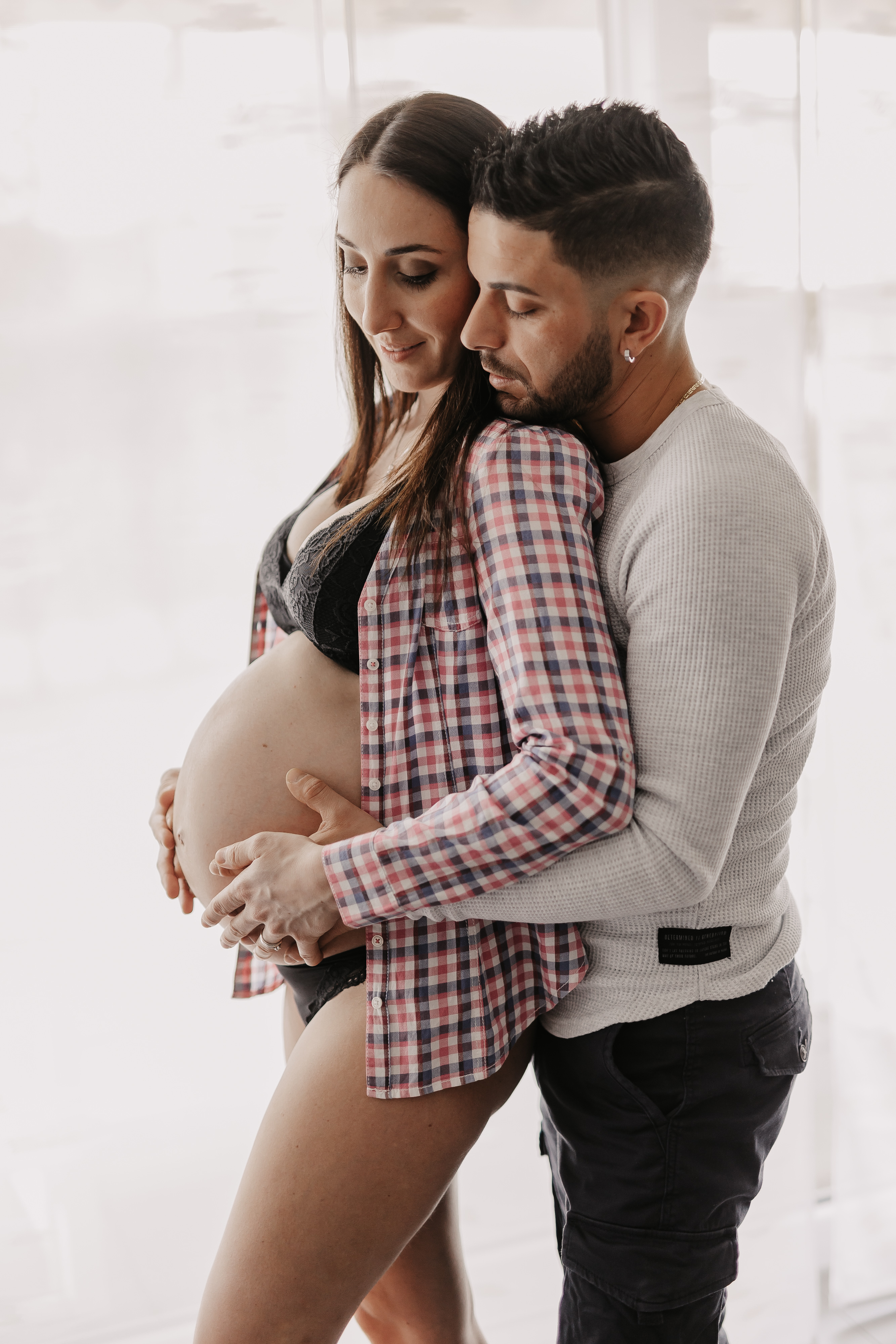 Eine schwangere Frau mit langen braunen Haaren, die ein kariertes Hemd und schwarze Unterwäsche trägt, wird von hinten von einem Mann mit kurzen dunklen Haaren und Bart umarmt, der ein weißes Langarmhemd und dunkle Hosen trägt. In dem sanft beleuchteten Raum teilen sie einen intimen Moment während ihres Babybauch-Fotoshootings.