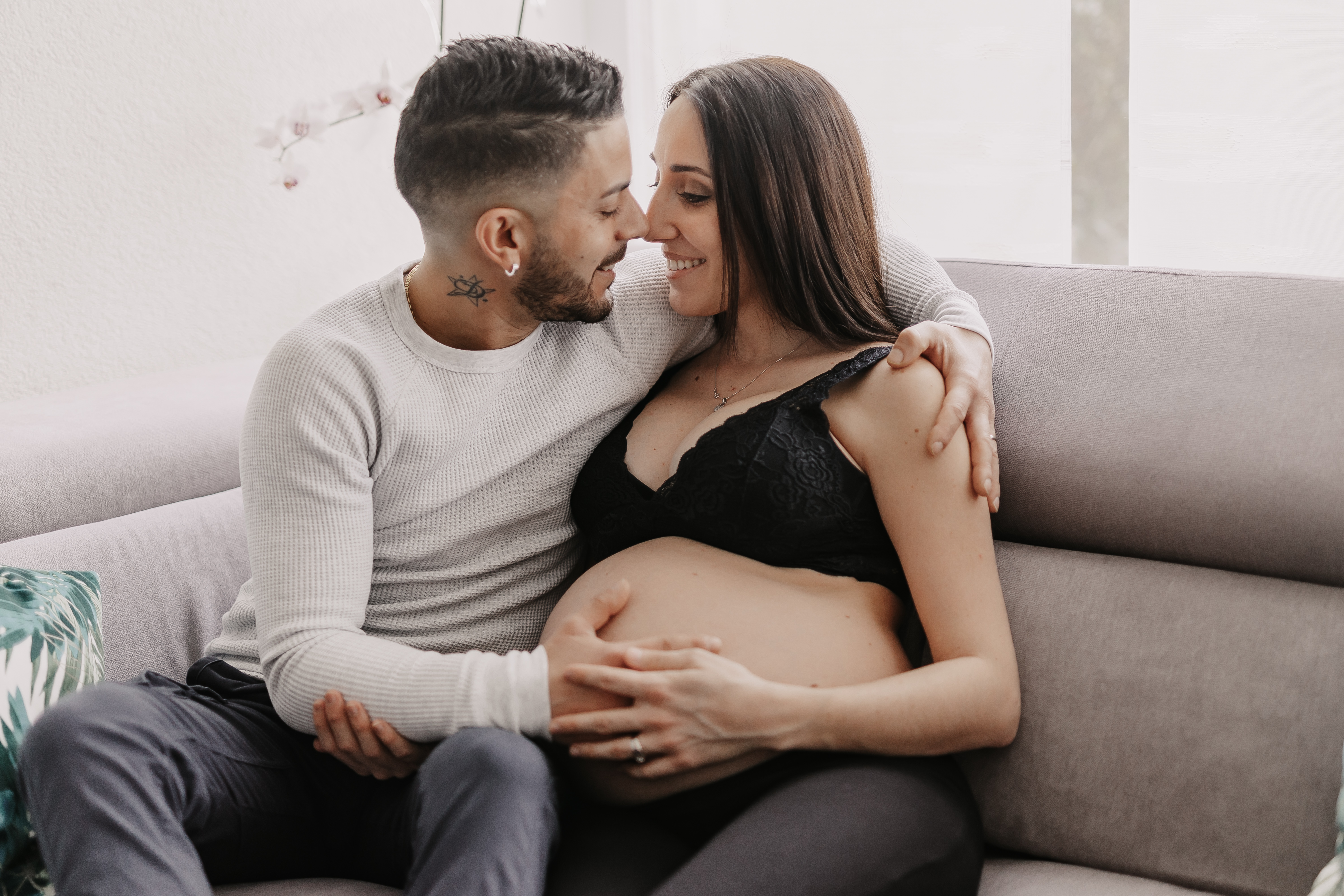 Eine schwangere Frau in einem schwarzen Oberteil sitzt auf einer Couch, berührt ihren Babybauch und lächelt einen Mann neben ihr an. Der Mann, der ein weißes Langarmshirt trägt, hält sie fest, während sie sich zu einem Kuss hinüberbeugen. Sie scheinen sich in Baden zu Hause zu fühlen, in einem lichtdurchfluteten Raum, der wie geschaffen ist für ein Schwangerschaftsshooting.
