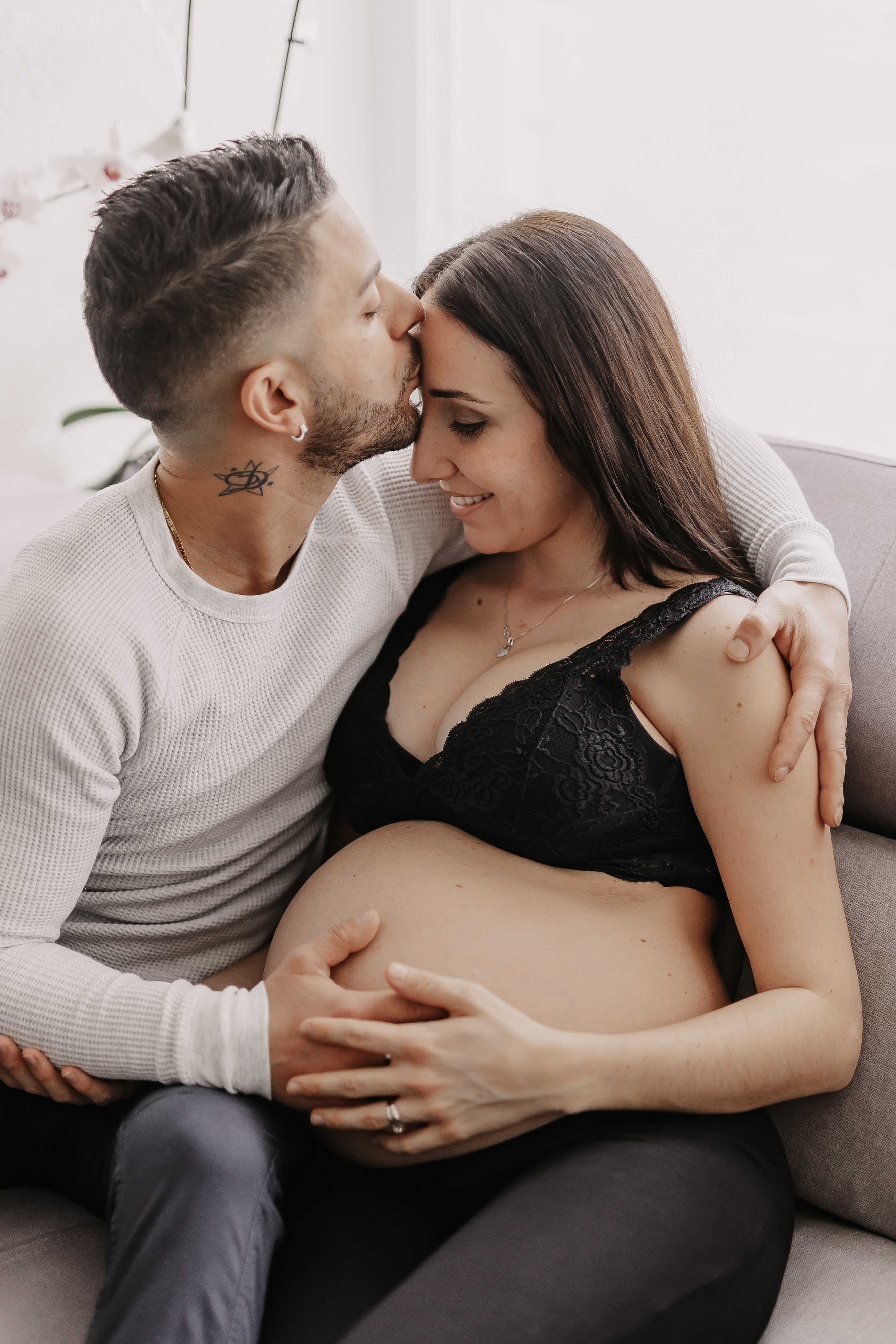 Ein Mann mit einem Tattoo am Hals küsst die Stirn einer lächelnden schwangeren Frau, während er sie auf einem Sofa umarmt. Die Frau, die einen schwarzen BH trägt, legt ihre Hand auf ihren Babybauch und beide wirken glücklich und liebevoll. Es ist der perfekte Moment für ein Schwangerschafts-Fotoshooting.