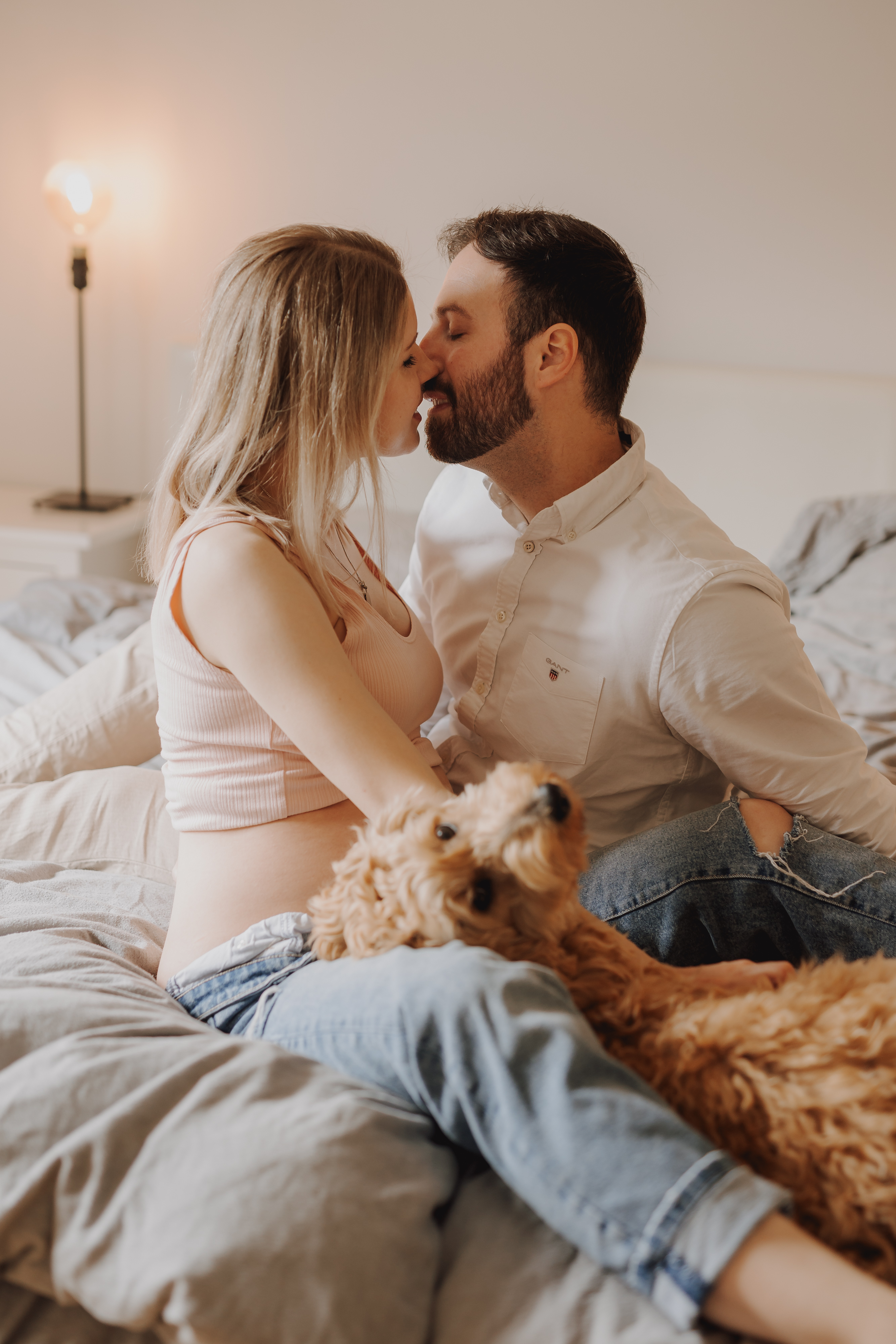 Ein Paar sitzt auf einem Bett und küsst sich während eines Babybauch-Fotoshootings. Die Frau mit sichtbarem Babybauch trägt ein rosa Top und Jeans, während der Mann ein weißes Hemd und Jeans trägt. Auf ihrem Schoß liegt ein flauschiger Hund, der in die Kamera blickt. Im Hintergrund leuchtet sanft eine brennende Lampe.