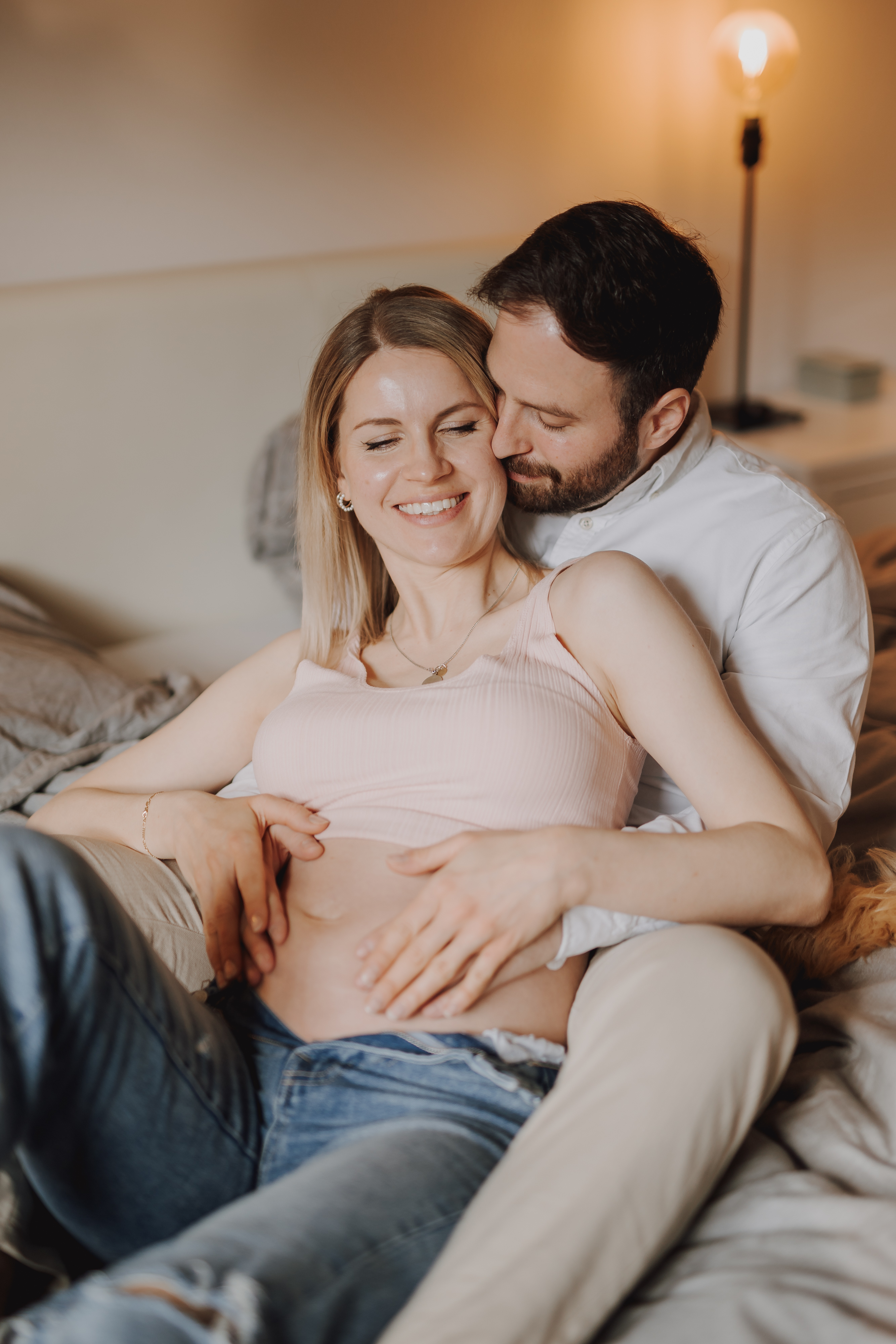 Ein Mann und eine Frau sitzen zusammen auf einem Bett. Die Frau, sichtlich schwanger, lehnt sich in die Umarmung des Mannes zurück. Beide lächeln und scheinen glücklich zu sein und genießen ihr intimes Schwangerschaftsshooting. Der Raum ist sanft beleuchtet, im Hintergrund eine warme Lampe.