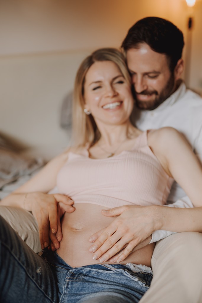 Eine schwangere Frau in einem rosa Tanktop und Jeans lächelt, während ein Mann hinter ihr sitzt, seinen Kopf auf ihre Schulter legt und ihren Babybauch mit beiden Händen hält. Sie sitzen auf einem Bett in einem warm beleuchteten Raum und fangen die Magie ihres Schwangerschafts-Fotoshootings zu Hause ein.