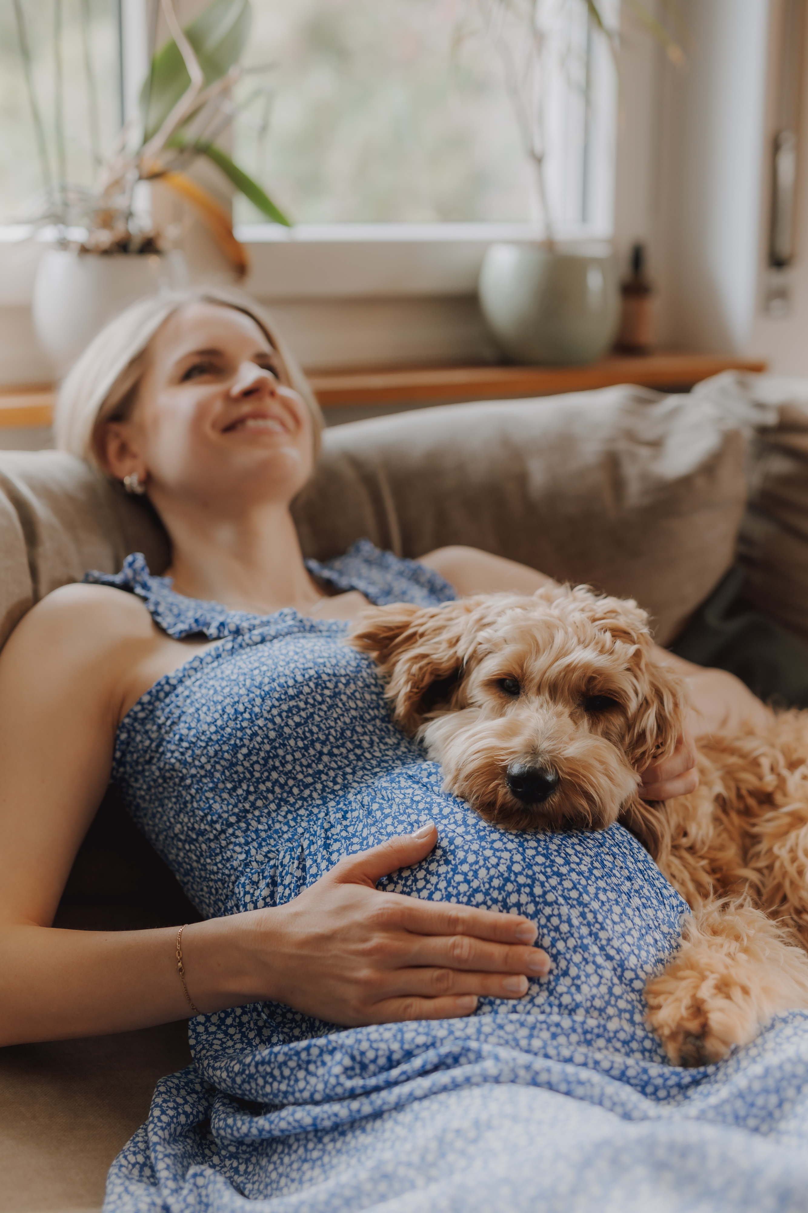 Eine schwangere Frau in einem blauen, geblümten Kleid liegt lächelnd auf einer Couch und hat ihre Hand auf ihrem Babybauch. Ein flauschiger, hellbrauner Hund kuschelt sich an ihren Bauch und beide wirken entspannt und zufrieden. Dieser heitere Zuhause-Moment ist perfekt für ein Fotoshooting. Im Hintergrund ist ein Fenster mit Topfpflanzen zu sehen.