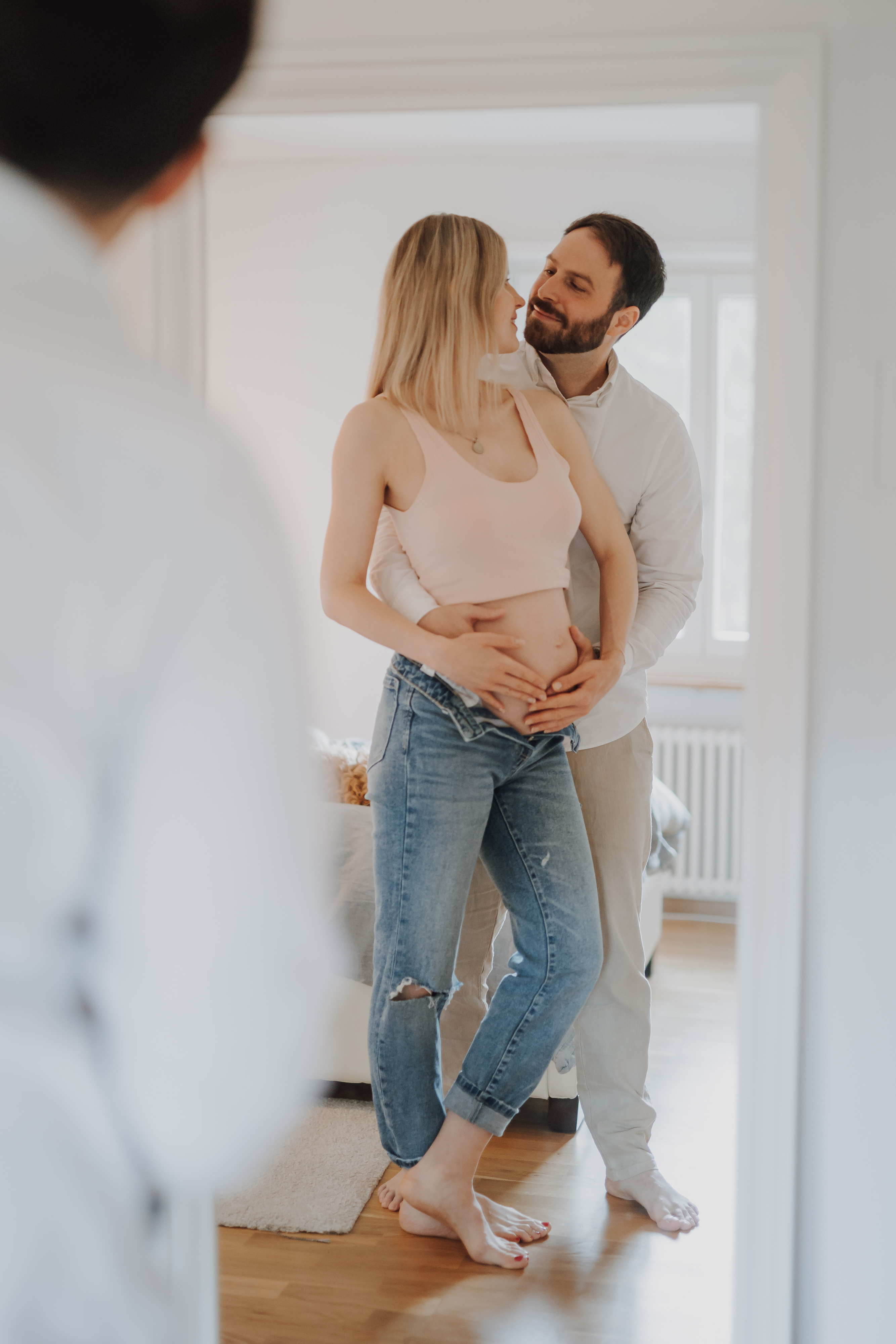 Eine schwangere Frau in legerer Kleidung steht in einem gemütlichen Raum, während ihr Partner sie von hinten umarmt. Beide lächeln herzlich. Eine weitere Person, leicht unscharf, steht im Vordergrund und blickt auf das Paar. Das natürliche Licht erfüllt die Szene und fängt einen perfekten Moment für ihr Schwangerschafts-Fotoshooting ein.