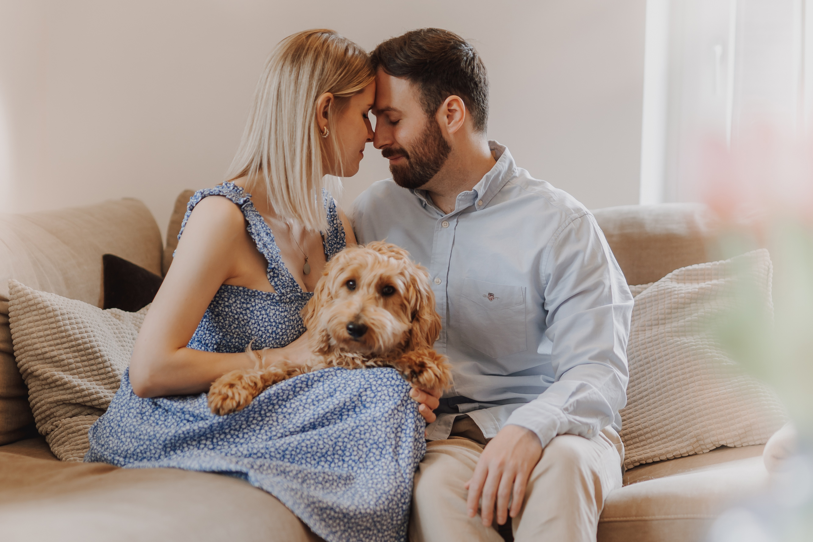 Ein Mann und eine Frau sitzen eng beieinander auf einer Couch und berühren sich während ihres Babybauch-Fotoshootings sanft mit den Stirnen. Die Frau trägt ein blaues Kleid und der Mann ein hellblaues Hemd, während sie liebevoll einen flauschigen braunen Hund auf dem Schoß halten. Der sanft beleuchtete Hintergrund verstärkt die gemütliche und intime Atmosphäre.